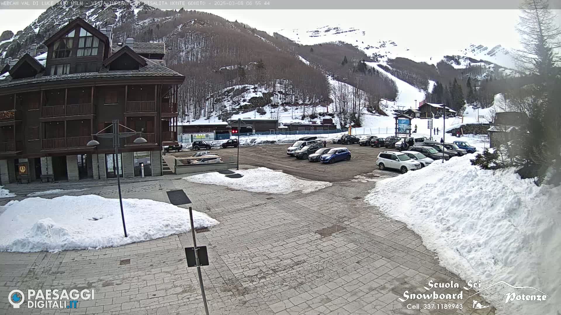 A snow-covered parking lot with cars sits in front of a multi-story brown building, with snow-covered mountains and ski slopes in the background.