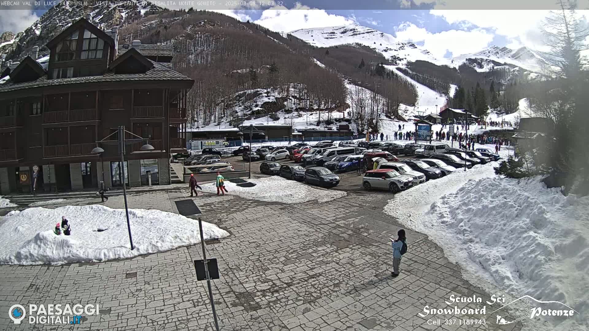 A parking lot with cars is situated in front of a multi-story building, with snow on the ground and in the background a snow-covered mountain under a partly sunny sky.