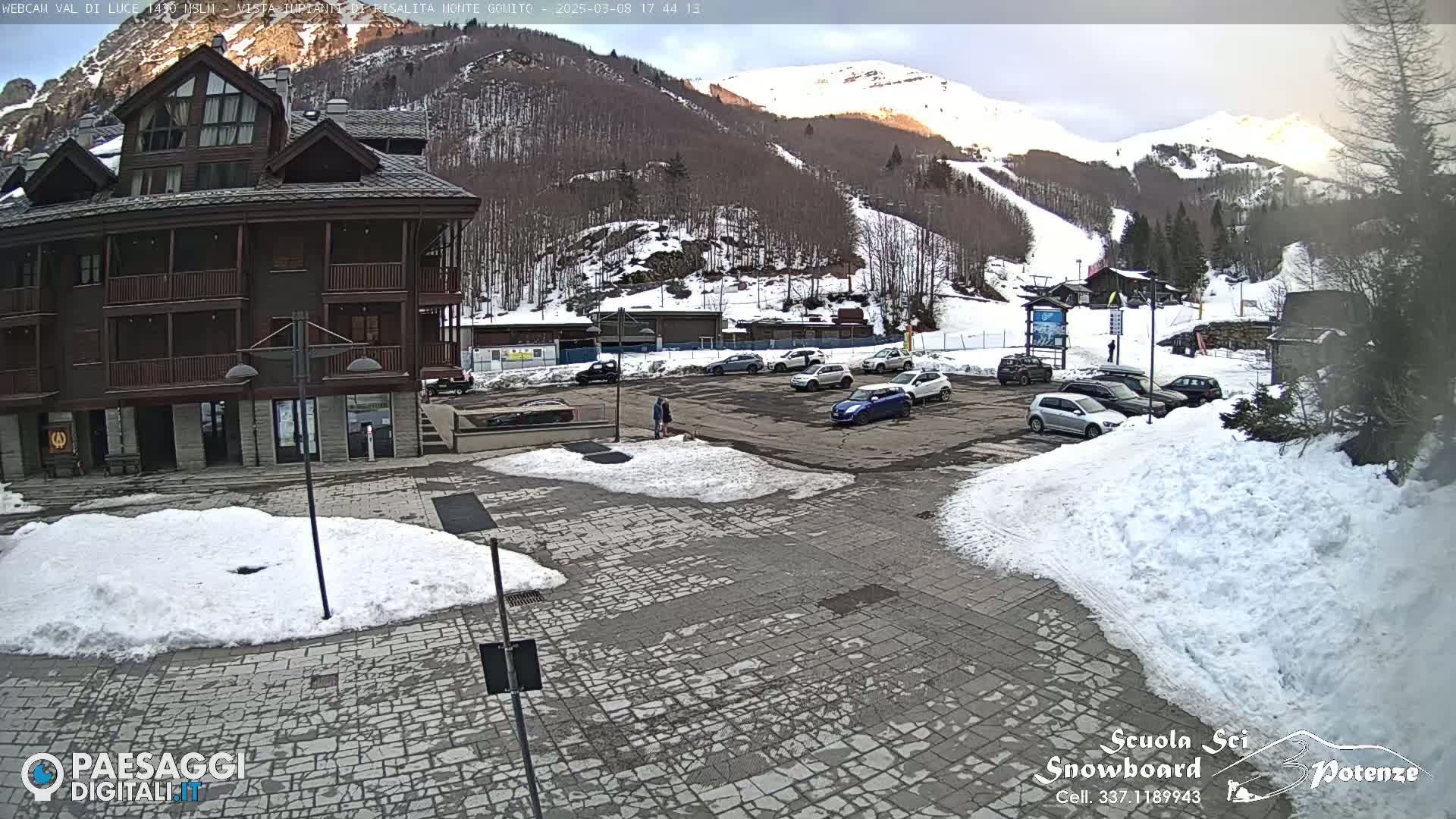 A parking lot with cars sits in front of a multi-story building, all covered in patches of snow, with a snowy mountainside and ski slopes in the background under a partly cloudy sky.