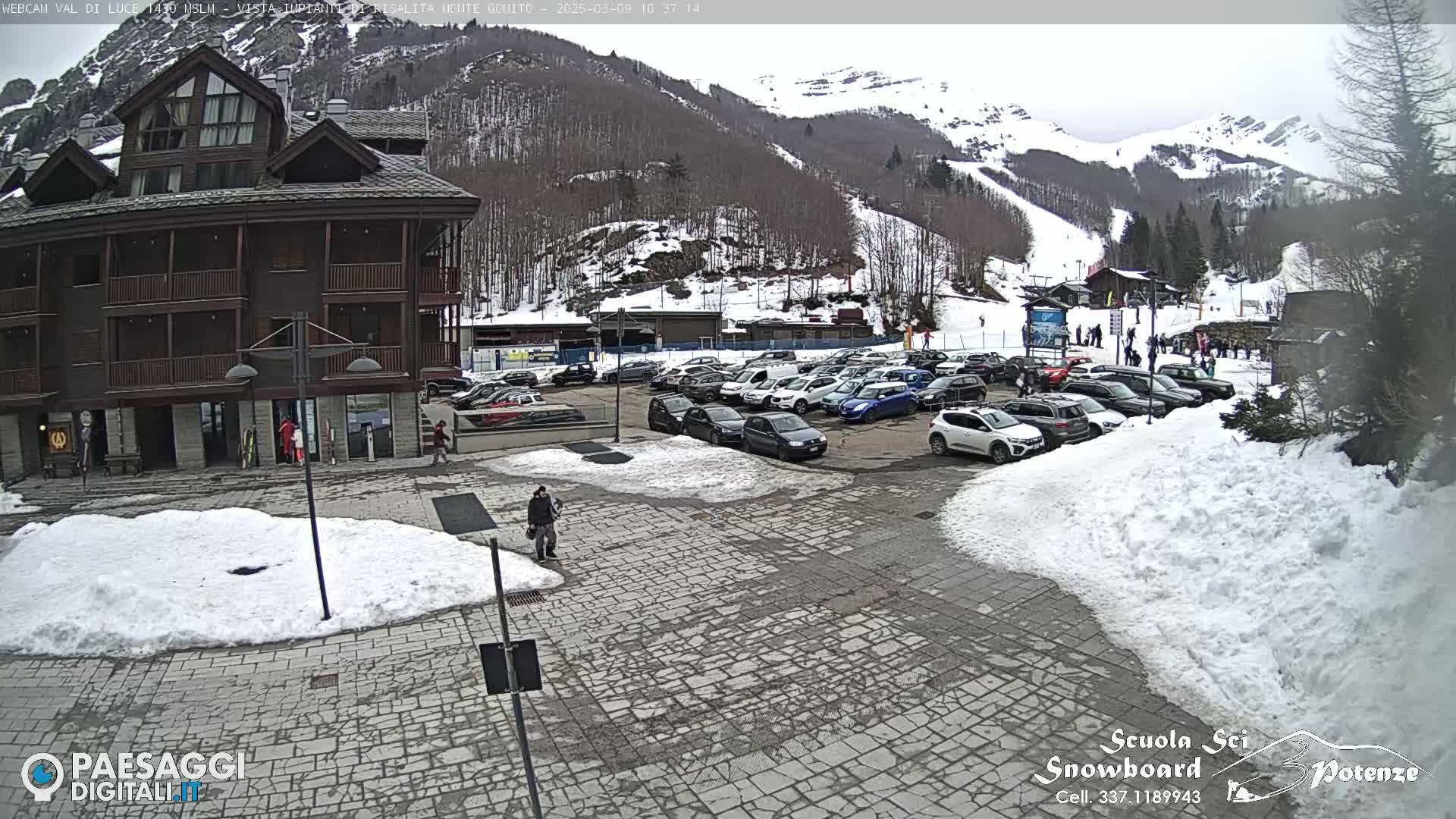 A parking lot filled with cars is situated in front of a brown multi-story building, with snow patches on the ground and a snow-covered mountainside in the background under an overcast sky.