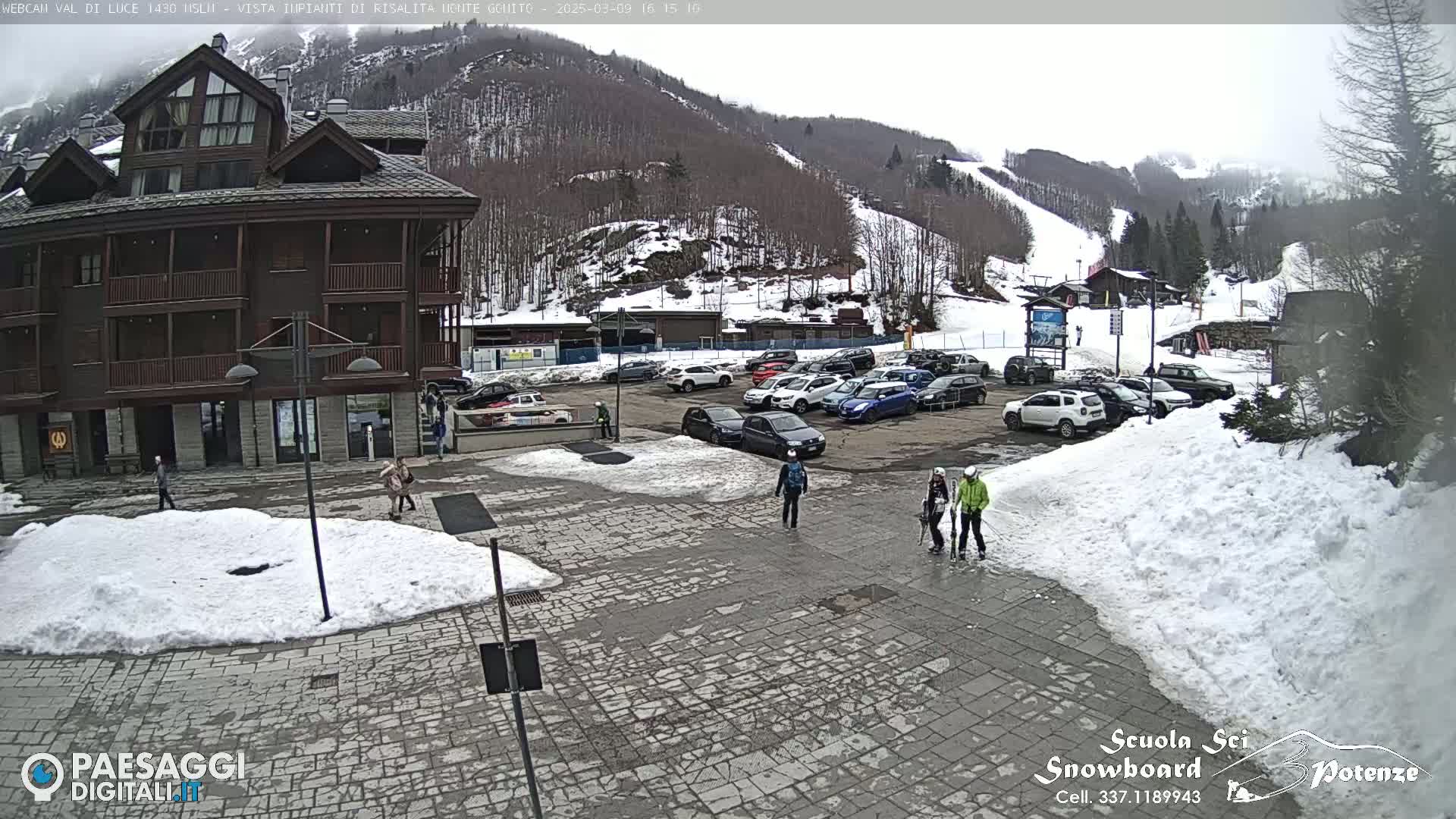 A stone plaza with several people walking, some cars parked in a lot, and a multi-story building sit in front of a snow-covered mountain with ski slopes, under an overcast sky.