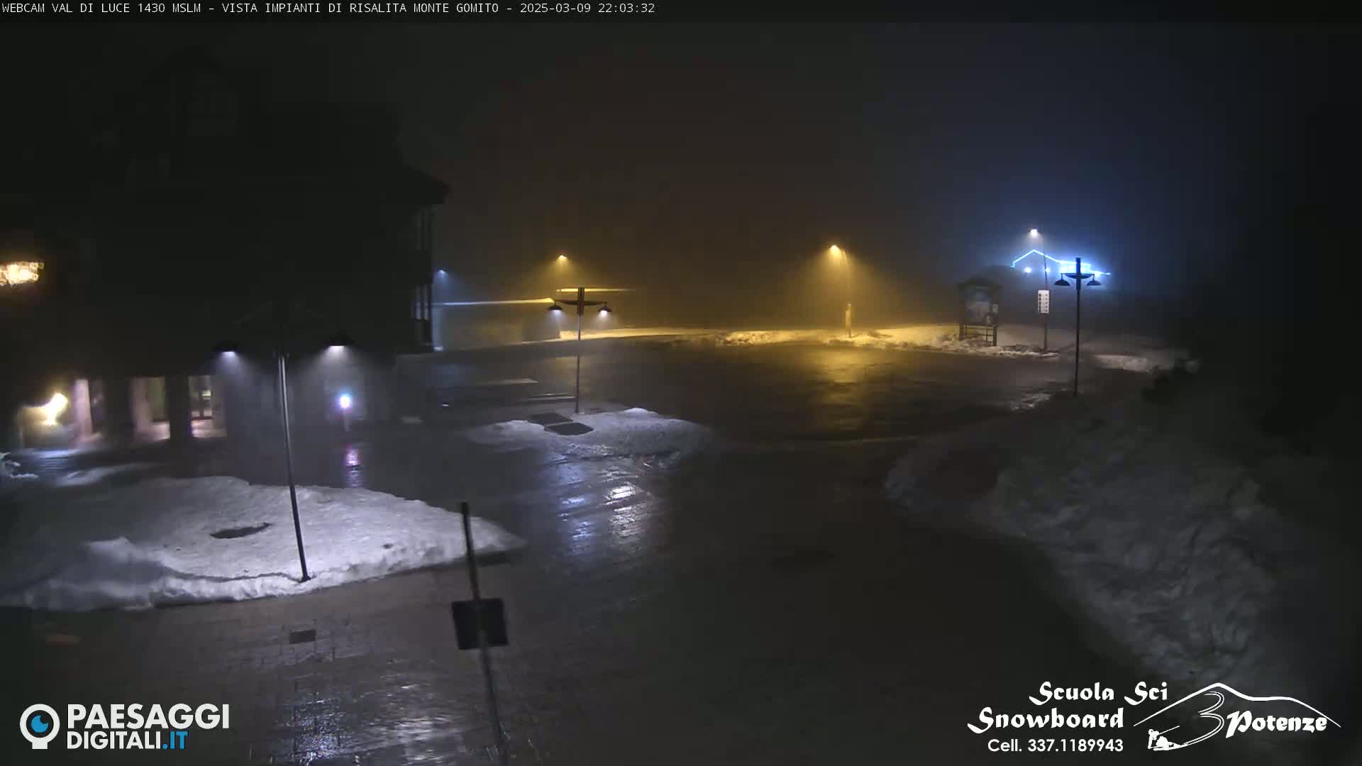 A foggy, nighttime scene shows a mostly empty parking lot with patches of snow, illuminated by streetlights, near a building.