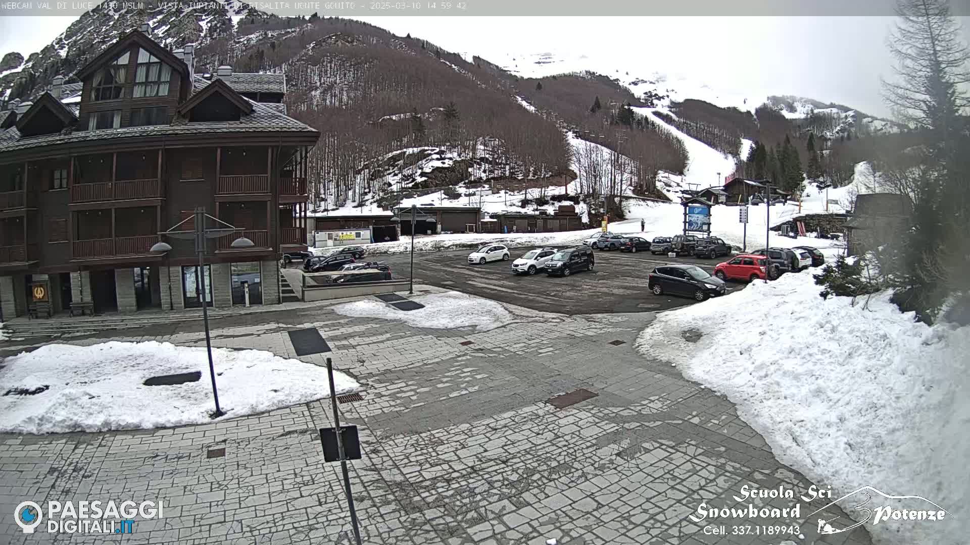 A snow-covered parking lot with cars and a multi-story wooden building is situated in front of a mountain with ski slopes, under an overcast sky.