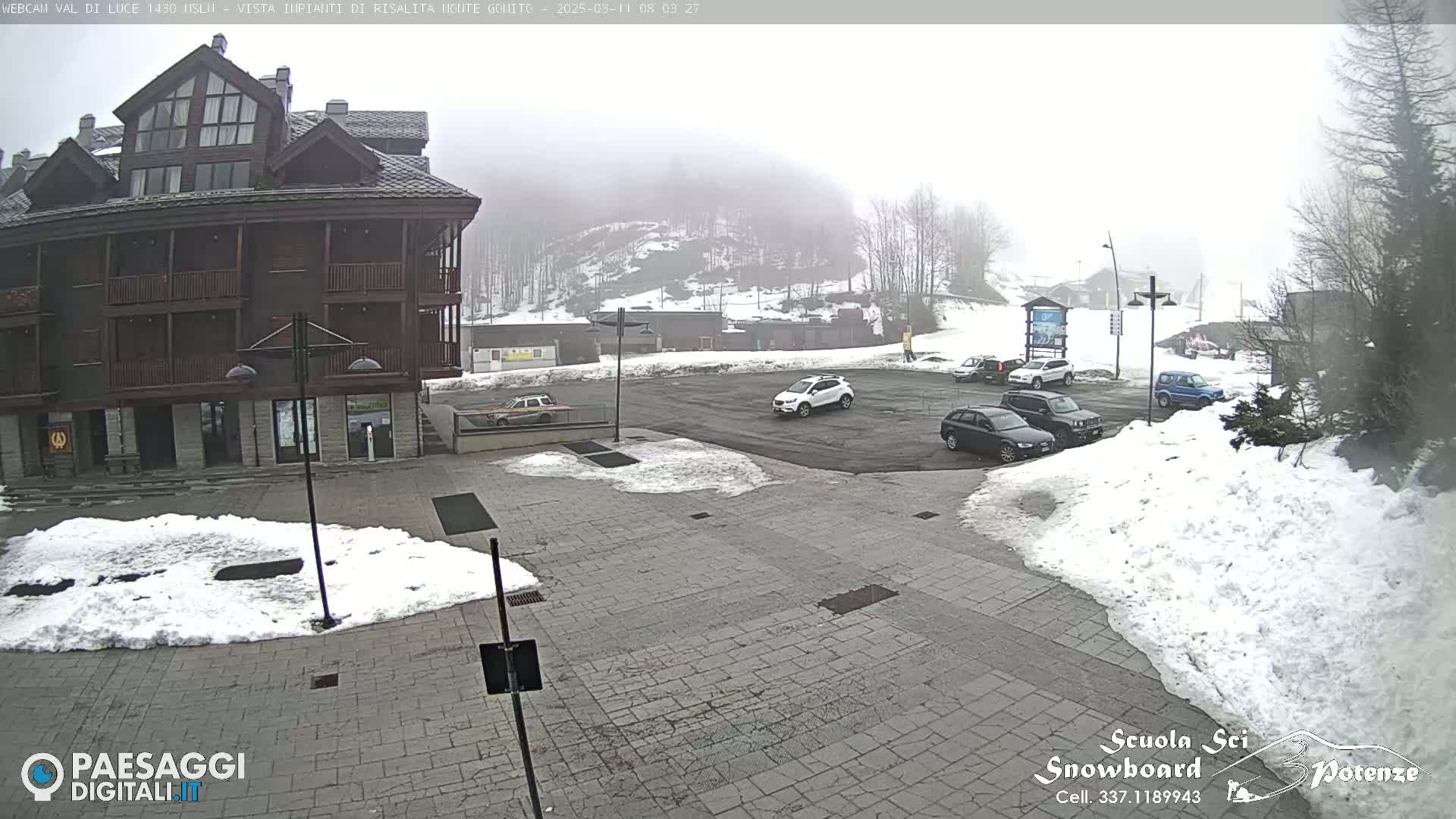 A mostly cloudy day shows a snow-covered parking lot with several cars in front of a multi-story wooden building, with a snowy hillside visible in the background.