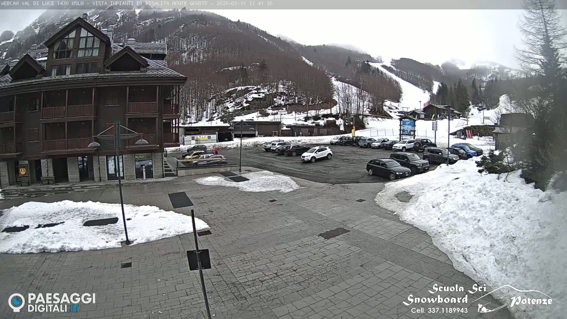 A parking lot with cars is in front of a multi-story building, with snow on the ground and a snow-covered mountain in the background under an overcast sky.