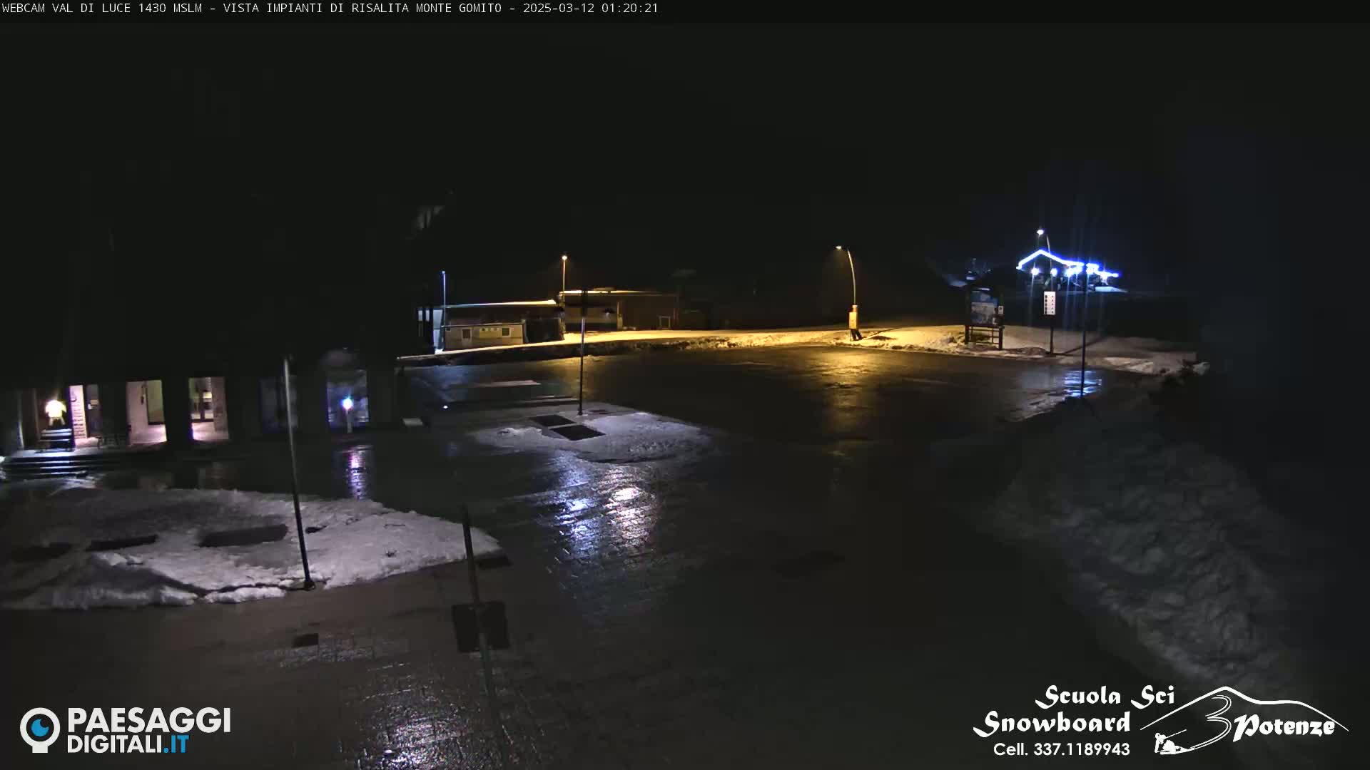 A nighttime view of a snow-covered parking lot and buildings illuminated by streetlights.