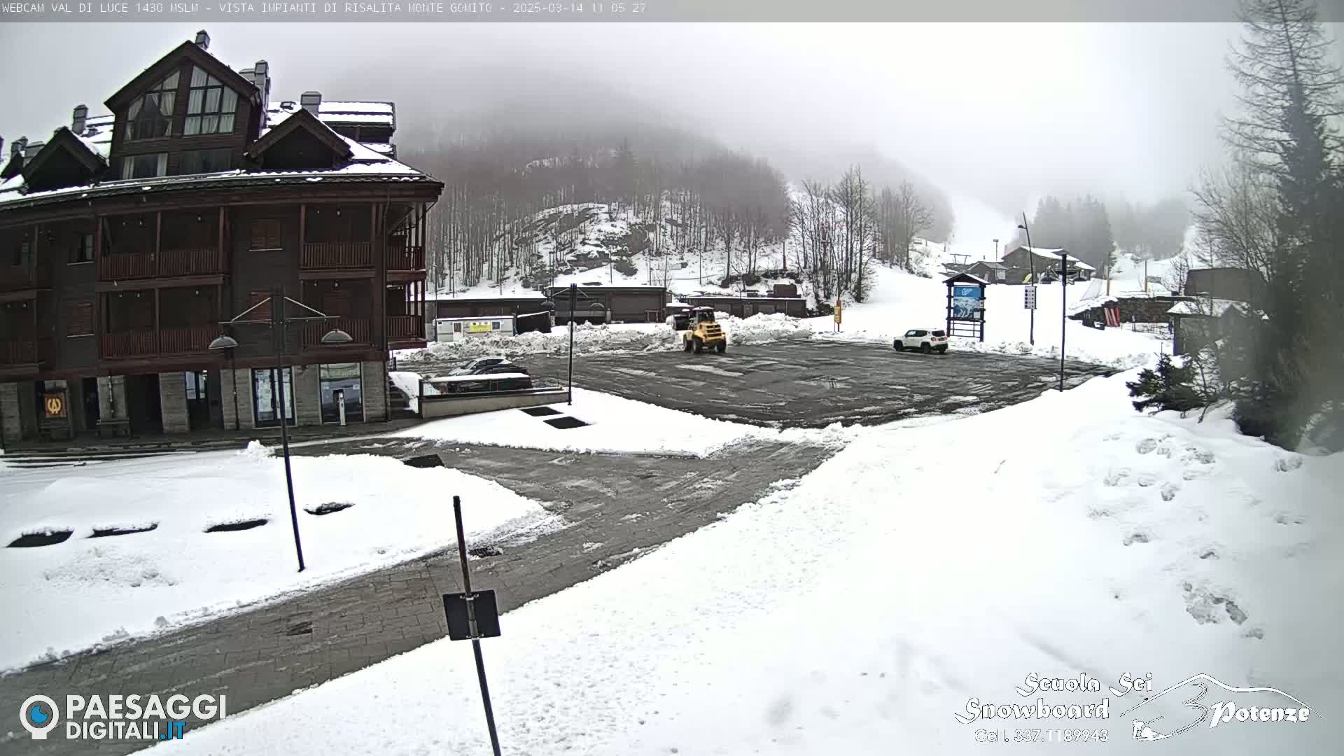 A snow-covered parking lot with a multi-story building and a small snowplow is visible in foggy weather conditions.