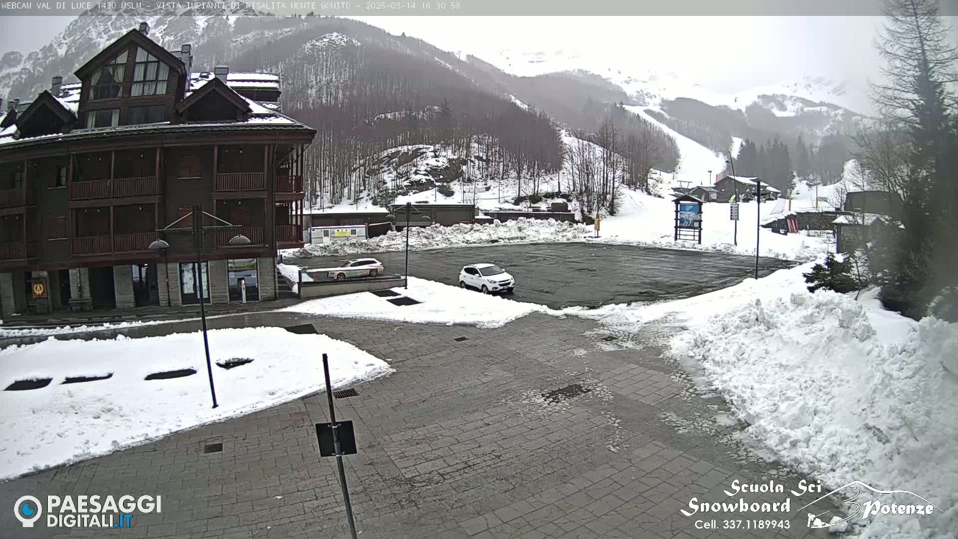 A snow-covered parking lot and paved area in front of a multi-story building, with a mountainous, snow-covered background.