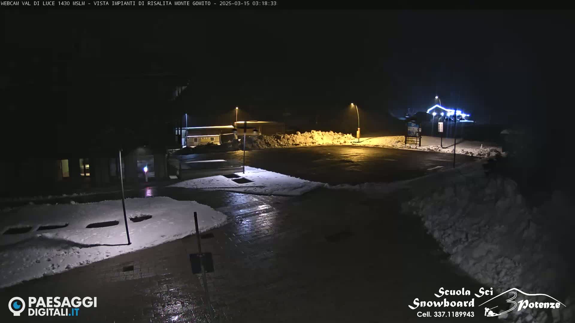 A nighttime, snow-covered parking lot is illuminated by streetlights, with snow piles visible nearby.