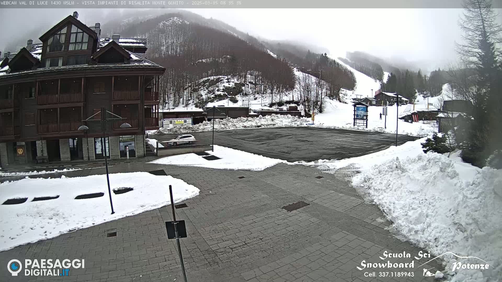 A snow-covered parking lot and paved area sit in front of a multi-story building, with a misty, snowy mountainside in the background.