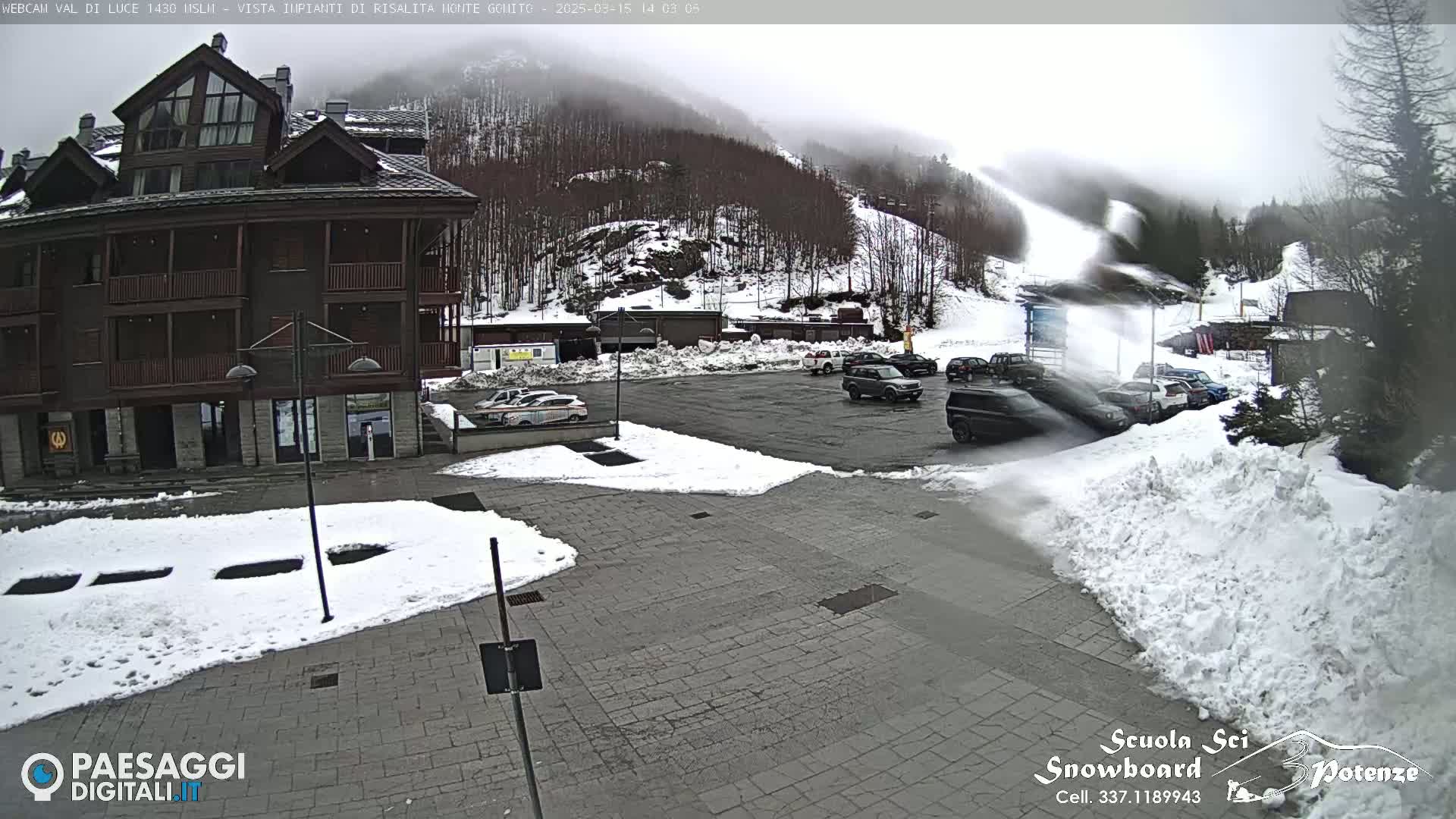 A snow-covered parking lot with several cars is in front of a multi-story building, with a snow-covered mountain and ski slope visible in the background under a cloudy sky.