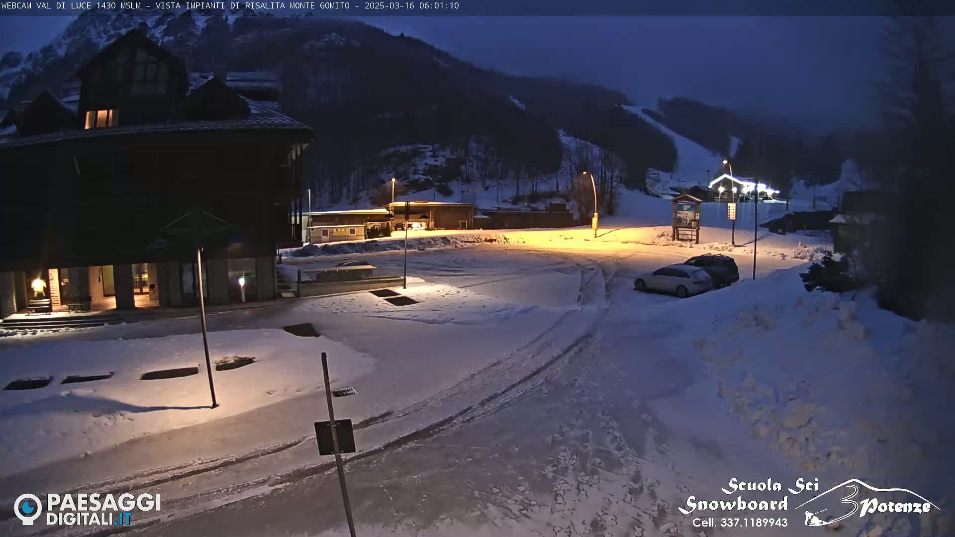 A snow-covered parking lot and building are illuminated at night, with a ski slope visible in the background under a dark sky.