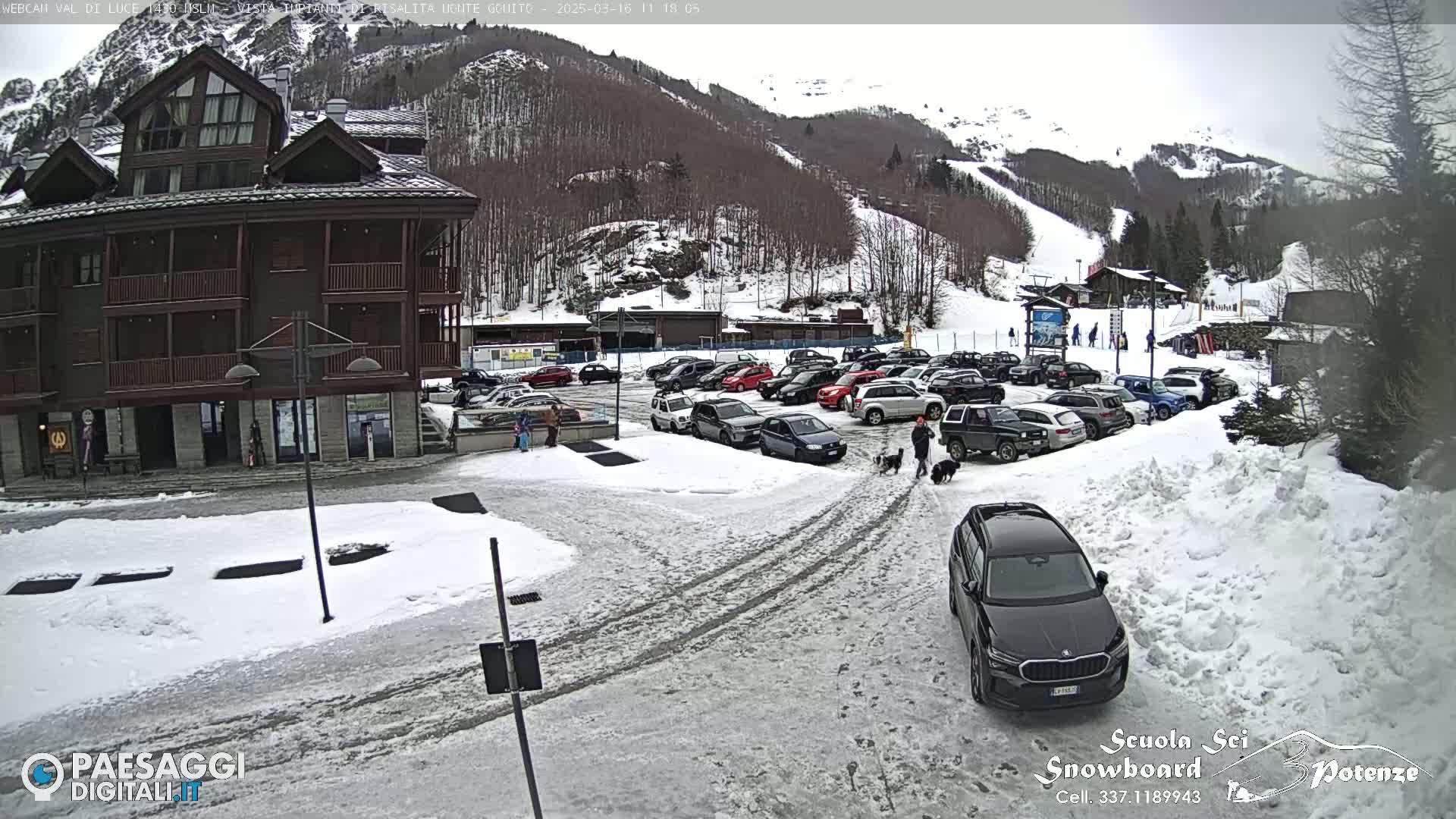 A snow-covered parking lot with cars and a multi-story building is situated near a snow-covered ski slope on a cloudy day.