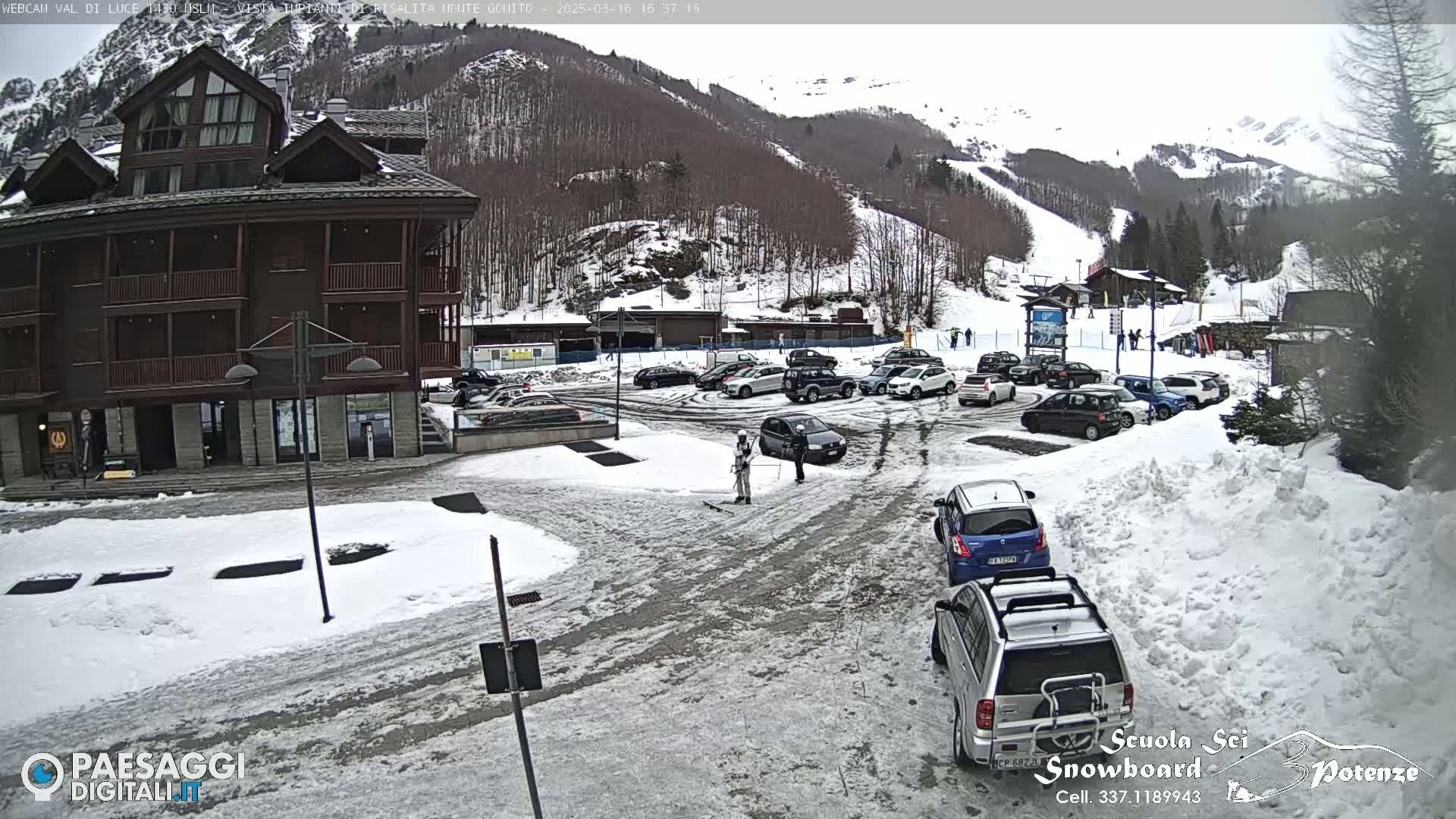 A snow-covered parking lot and street scene in front of a multi-story building, with cars parked and a few people walking nearby, against a background of a snowy mountain slope with ski runs.