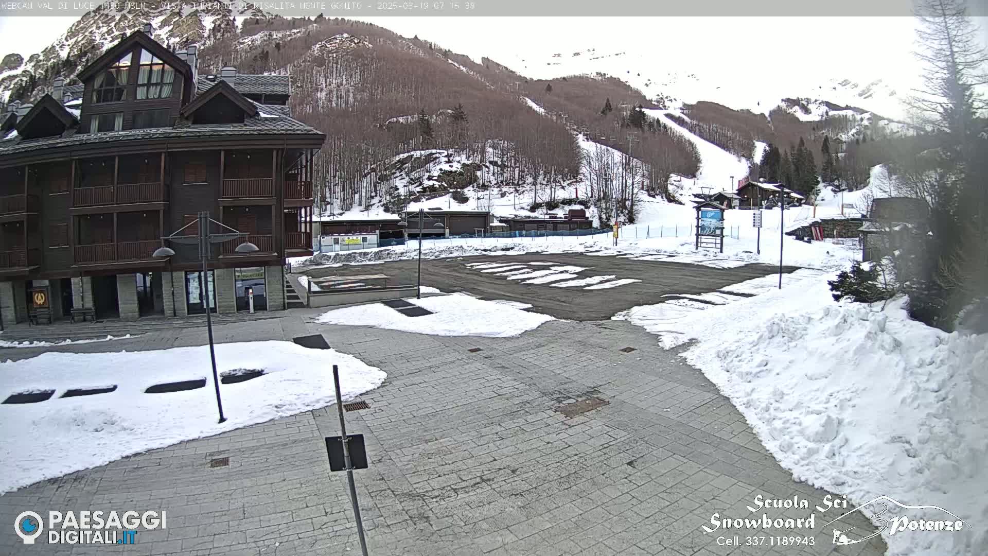 A snow-covered plaza sits in front of a dark brown multi-story building with a mountain and ski slope in the background under a cloudy sky.