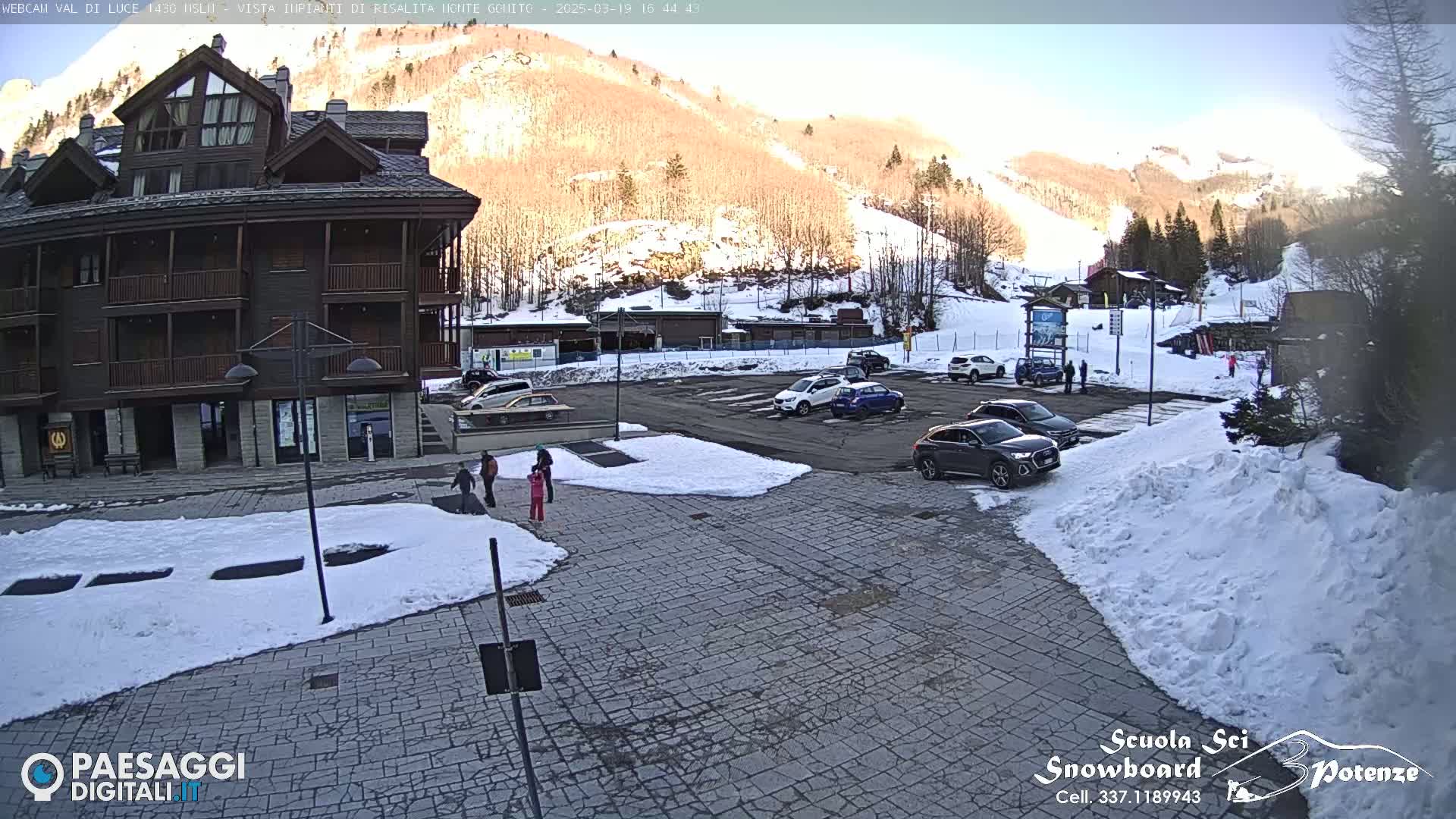 A multi-story wood-sided building sits in a parking lot partially covered in snow, with a sunny mountainside in the background.