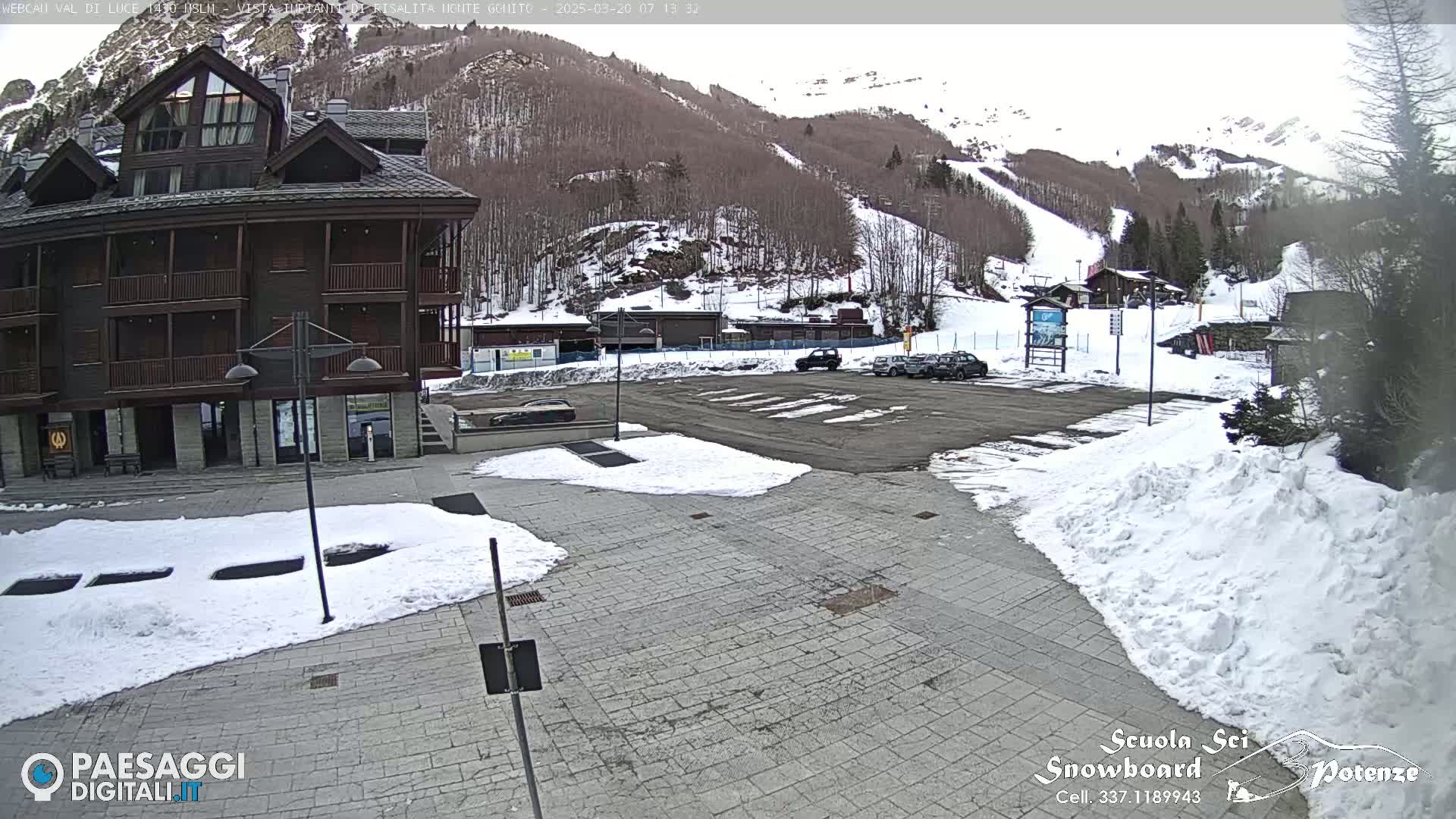 A snow-covered paved area with a multi-story building and a parking lot in the foreground, overlooks a ski slope on a partly cloudy day.