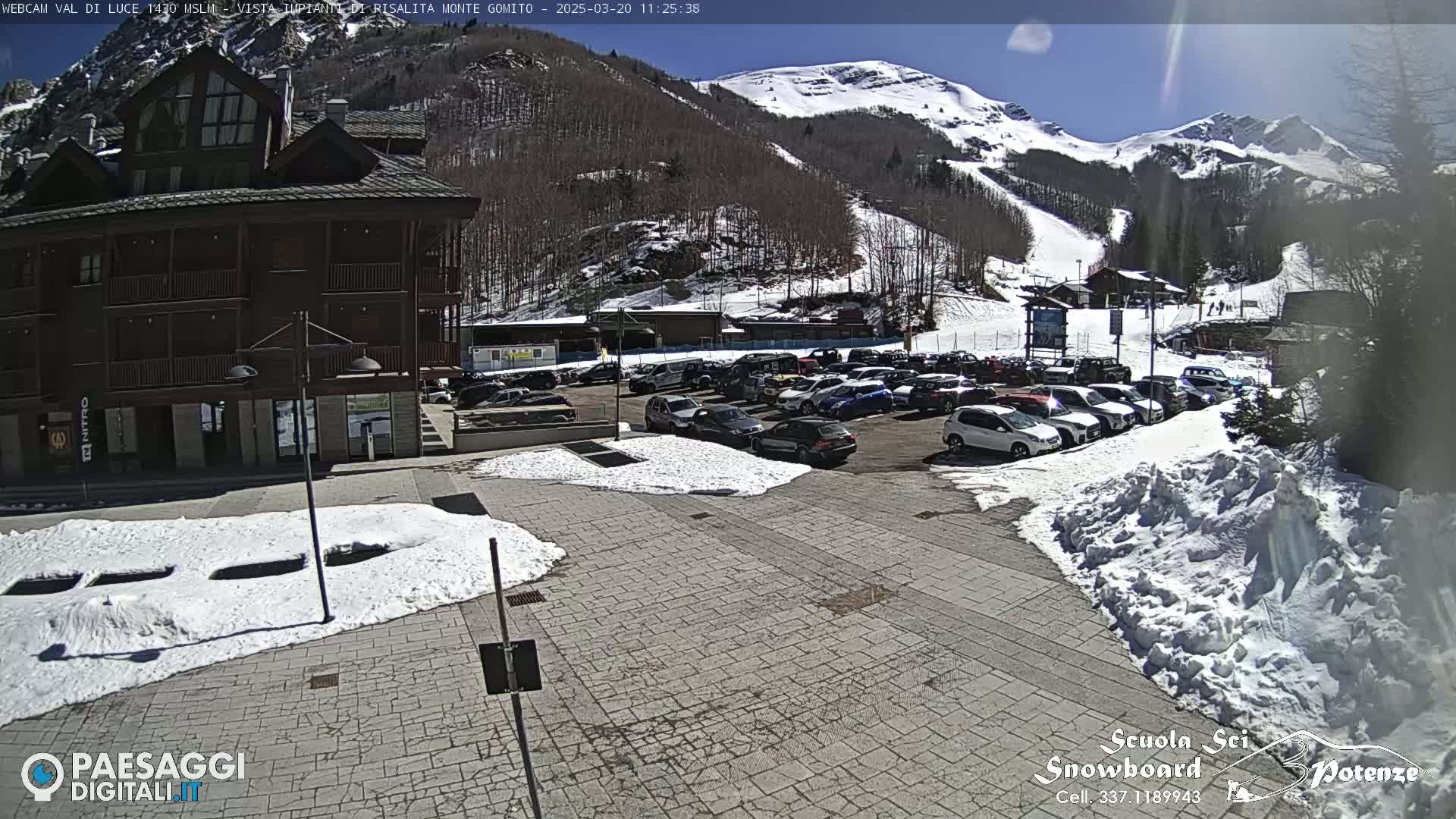 A sunny day reveals a parking lot full of cars in front of a multi-story building near a snow-covered ski slope and mountain.
