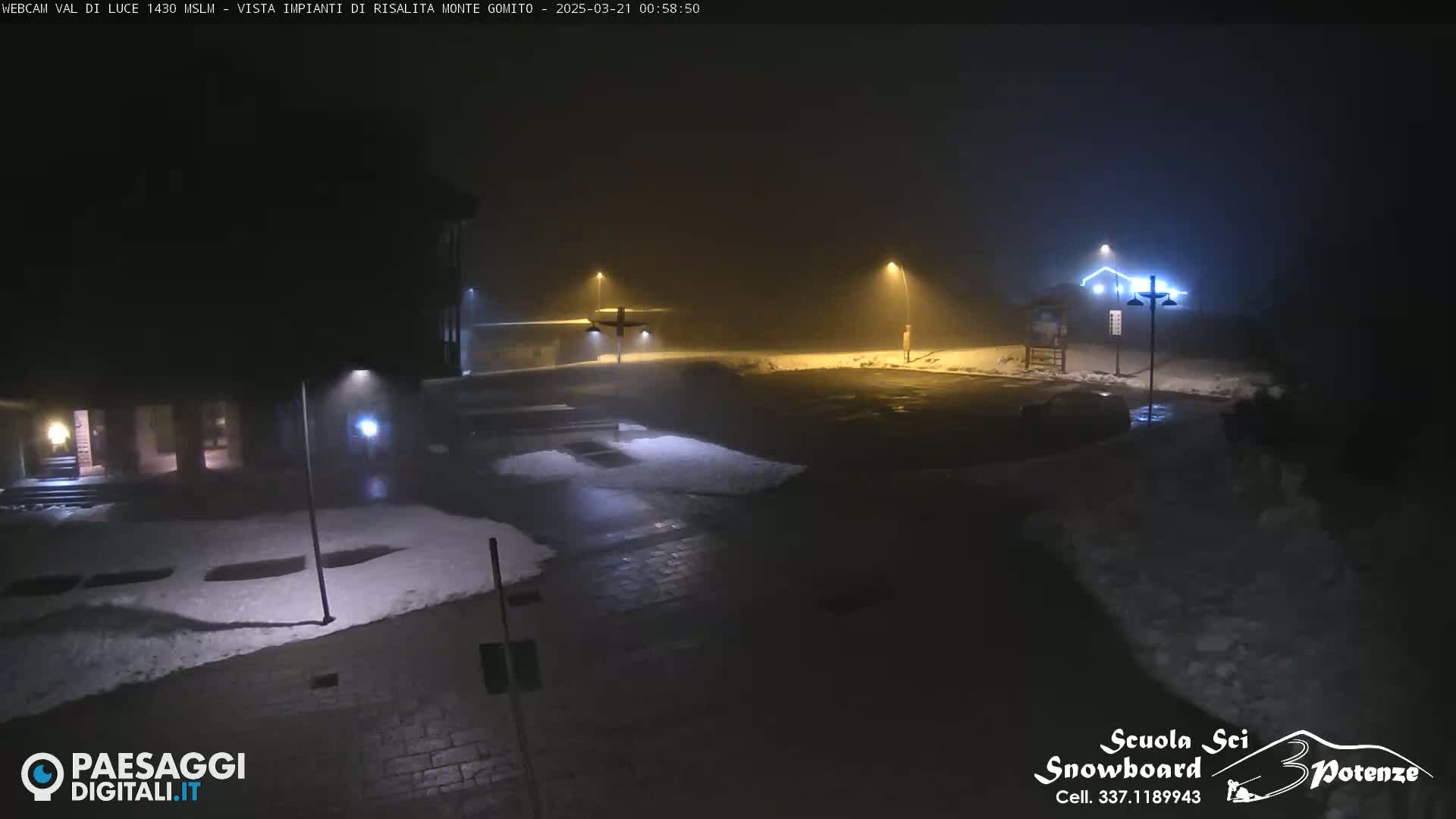 A foggy, snowy night scene shows a mostly empty parking lot illuminated by streetlights near a building.