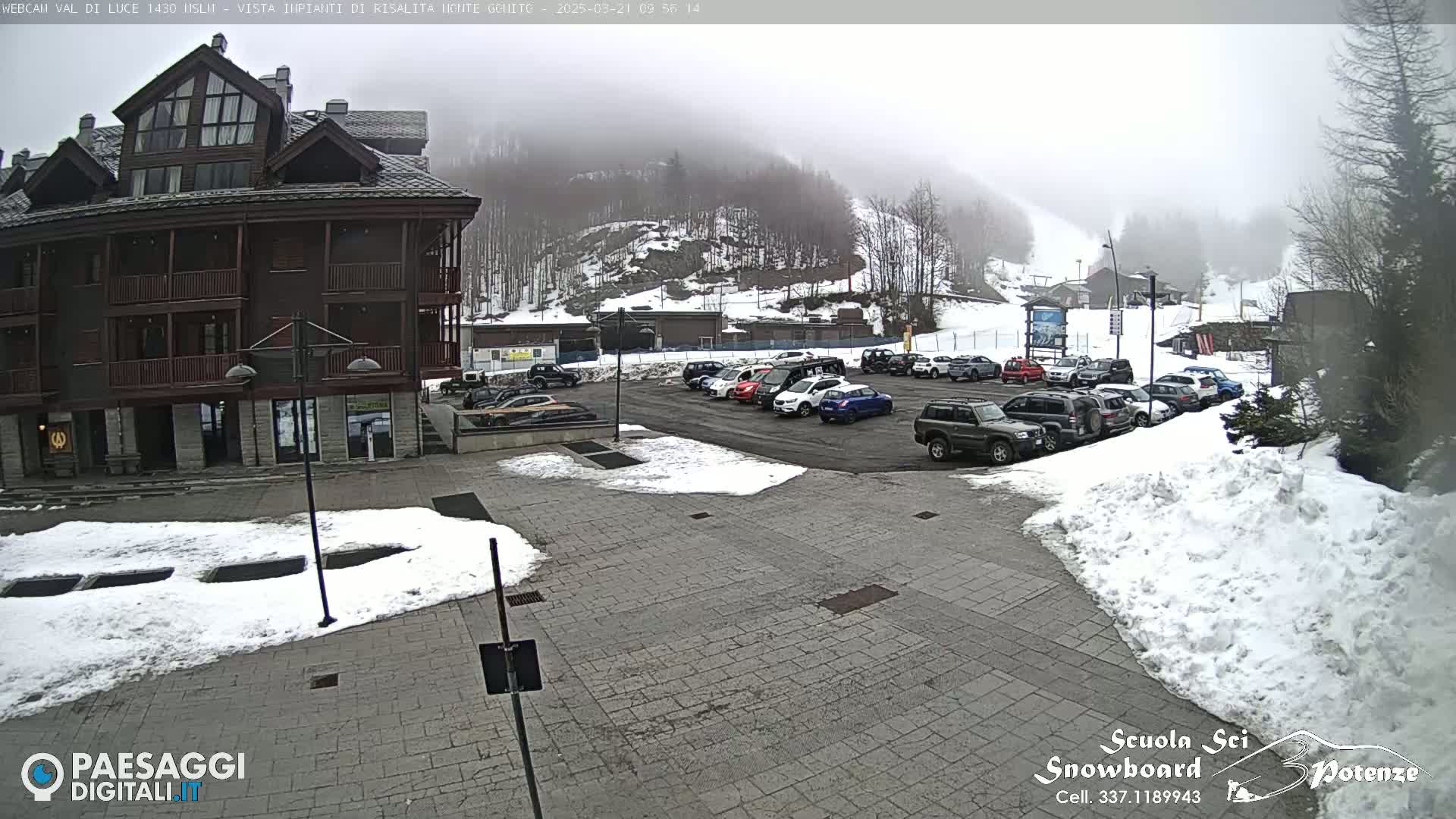 A foggy, snowy scene shows a parking lot full of cars in front of a multi-story building, with a snow-covered plaza in the foreground and a snow-covered hillside in the background.