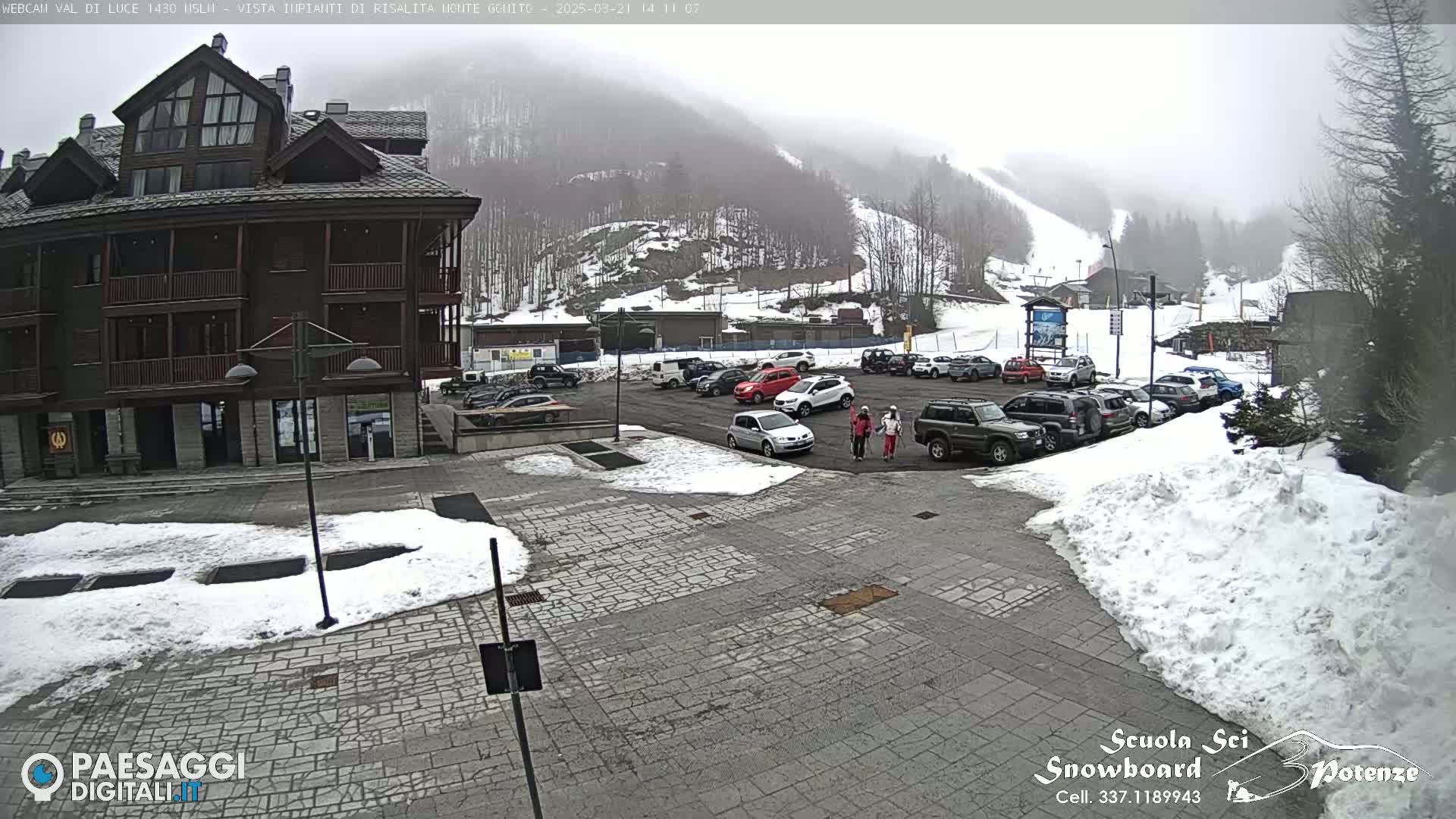 A snow-covered parking lot with cars and a multi-story building is visible, with a foggy mountain background showing a ski slope.