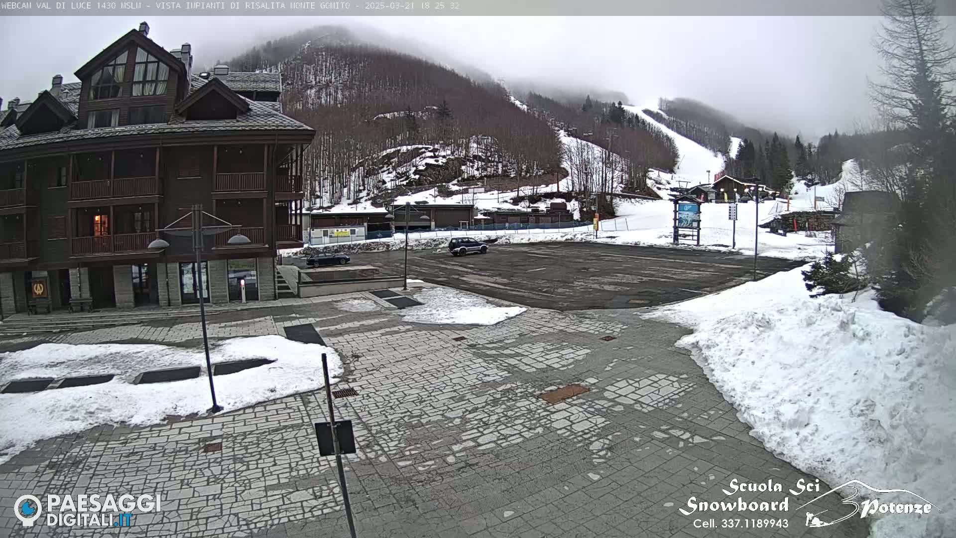 A snow-covered parking lot and a multi-story wooden building are visible, with a partially cloudy and hazy sky overlooking a snow-covered mountainside with ski slopes.