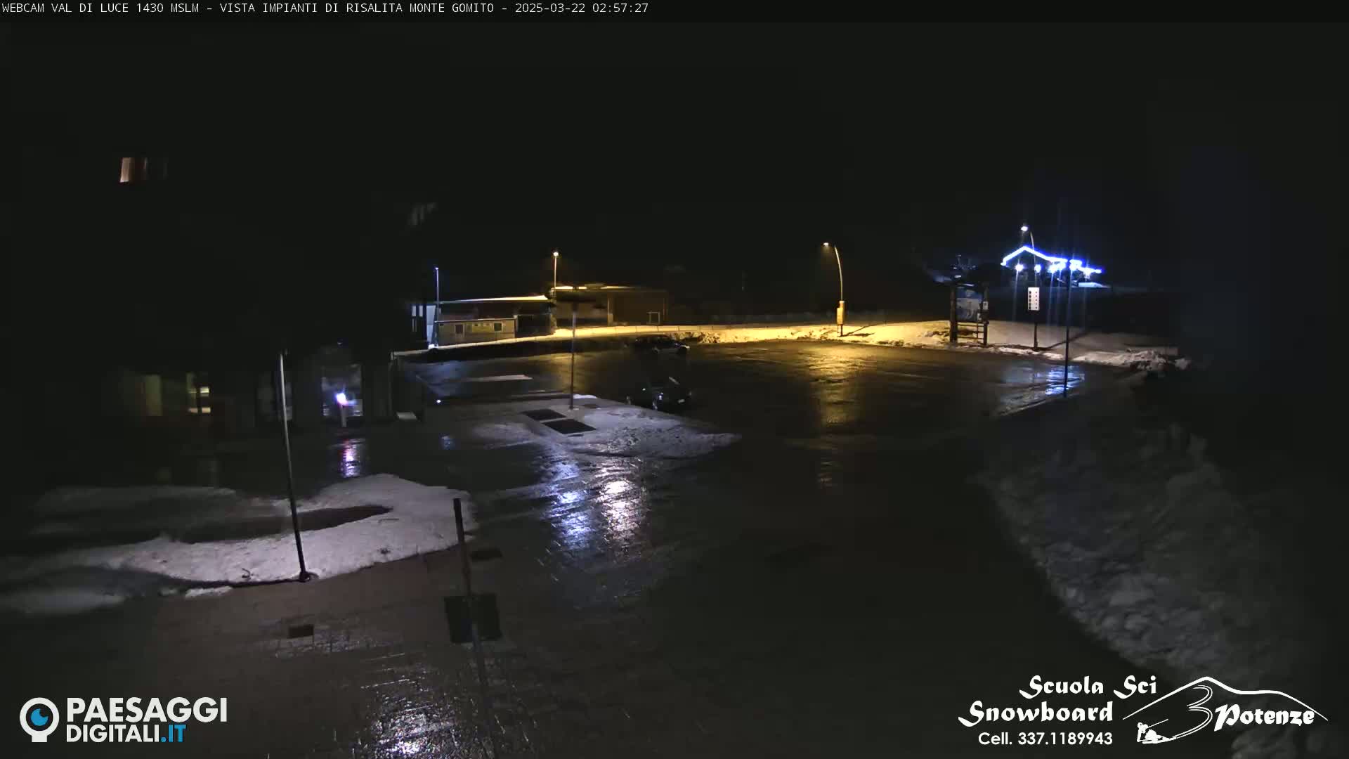A nighttime, snow-dusted parking lot is illuminated by streetlights, with a few cars visible and some small buildings in the background.