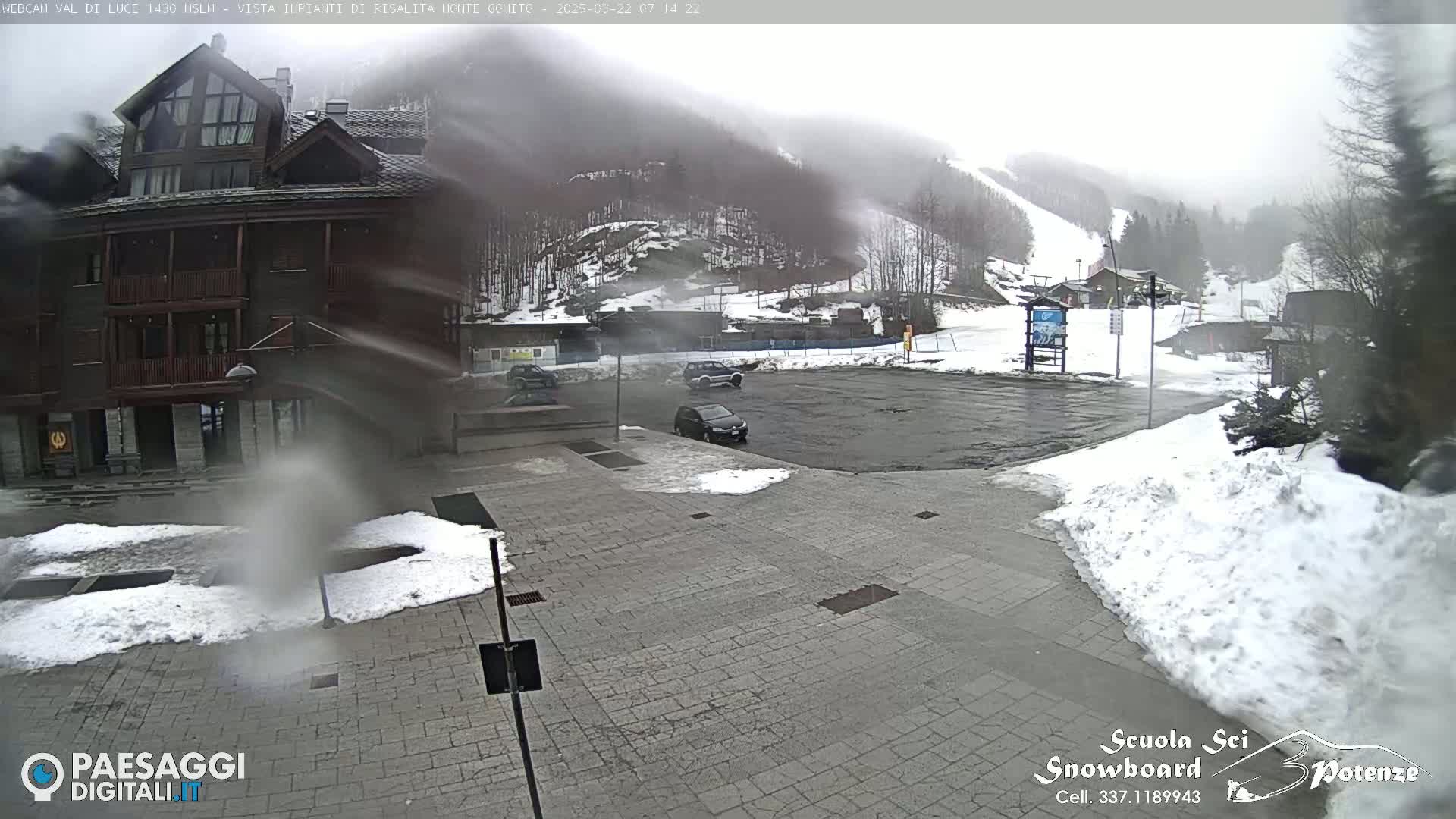 A mostly empty paved parking lot with a few cars is partially covered in snow, in front of a multi-story building and a snow-covered mountain with ski slopes visible in the hazy, snowy weather.