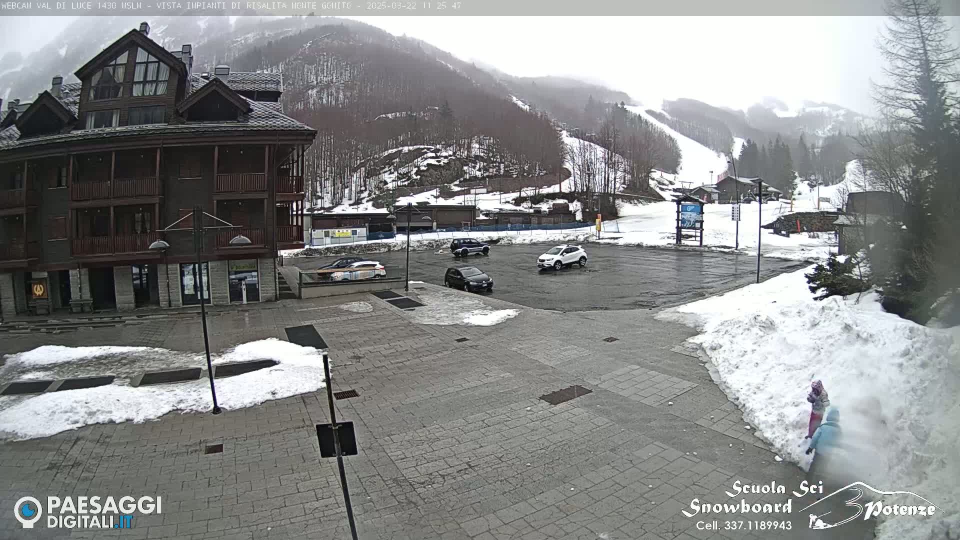 A snow-covered parking lot with several cars and a multi-story building is seen in front of a snow-covered mountainside with ski slopes.