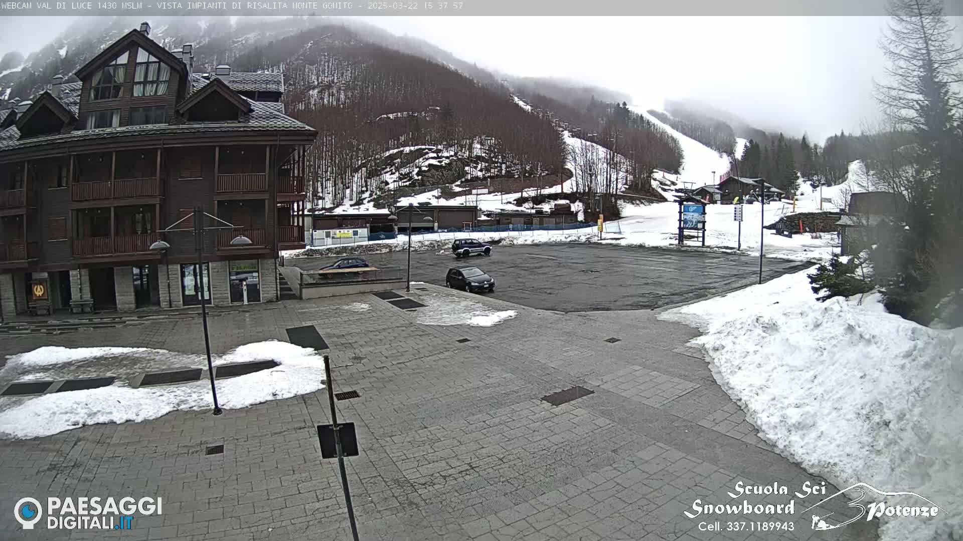 A paved plaza with patches of snow, a few parked cars, and a multi-story brown building is situated in front of a snow-covered mountain with ski slopes under an overcast sky.