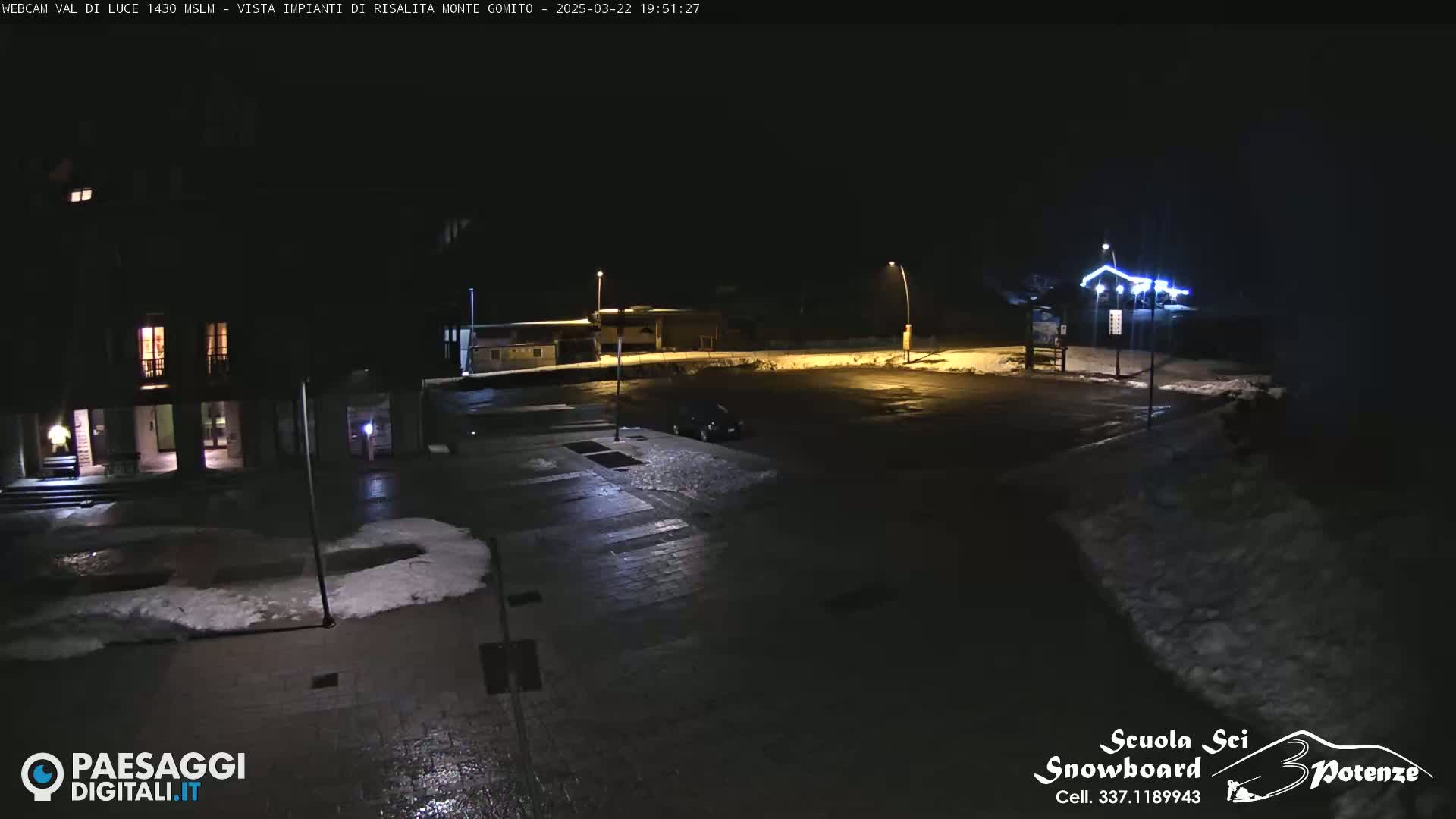A nighttime view of a mostly empty parking lot with patches of snow, illuminated by streetlights, showing a building with lights on in the background.