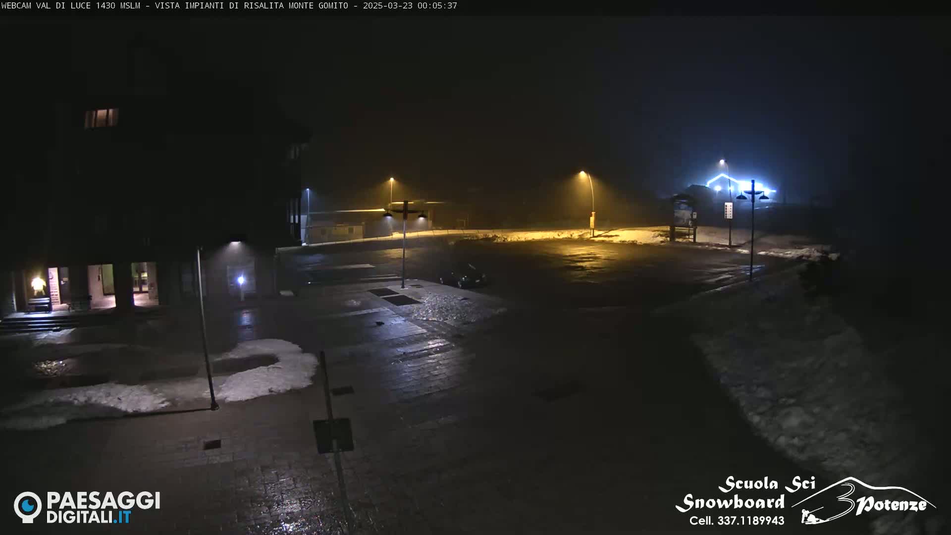 A foggy, dark nighttime scene shows a mostly empty paved area with patches of snow, illuminated by streetlights, near a building.