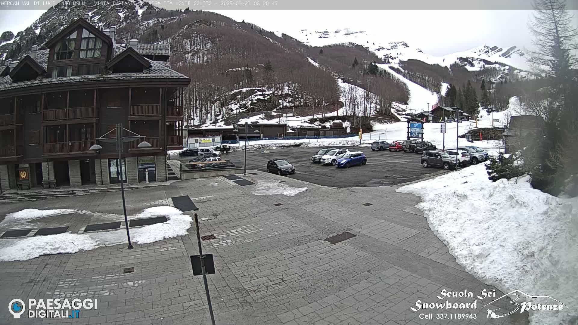 A paved plaza with patches of snow, several cars parked in a lot, and a multi-story building in the background against a snow-dusted mountainside.