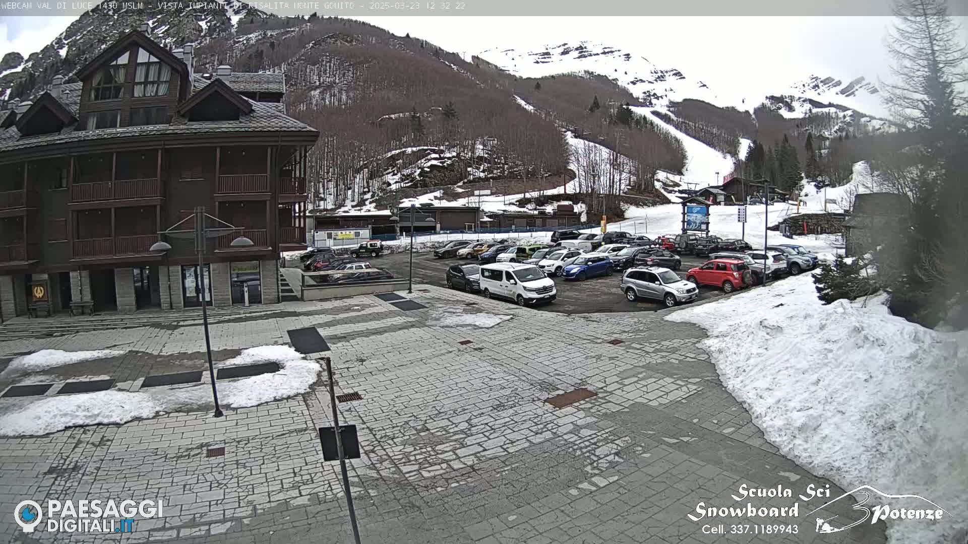 A parking lot with cars is in front of a multi-story building, with snow on the ground and a snowy mountainside in the background.