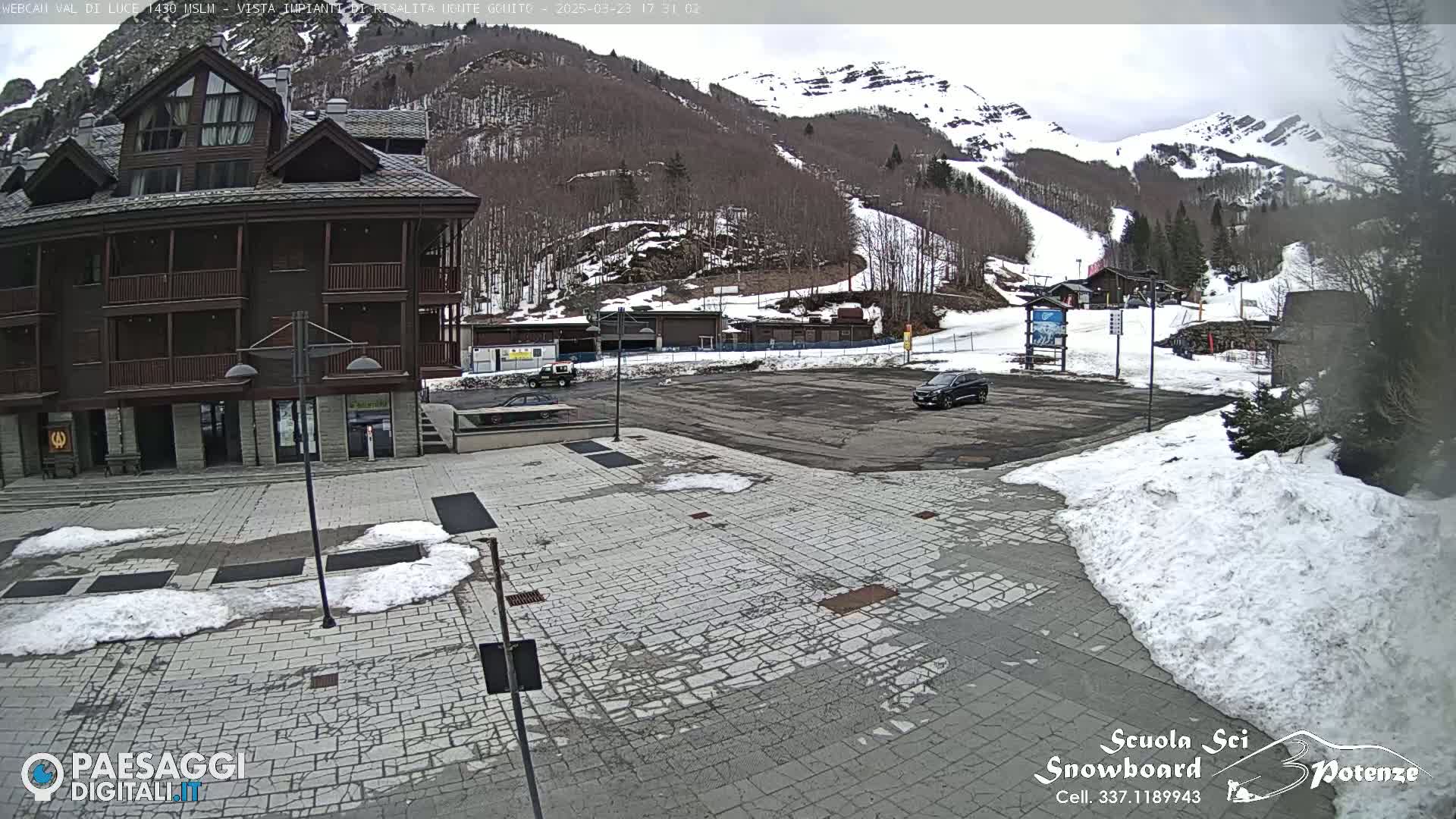 A paved plaza with patches of snow is situated in front of a multi-story building, with a parking lot and snowy mountains in the background under an overcast sky.