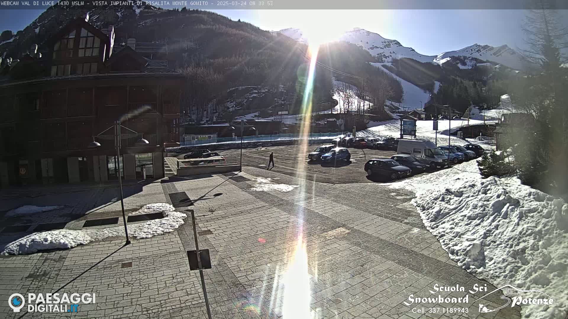 A sunny day in a snow-covered mountain resort town shows a parking lot with cars, a building, and patches of snow on the ground.