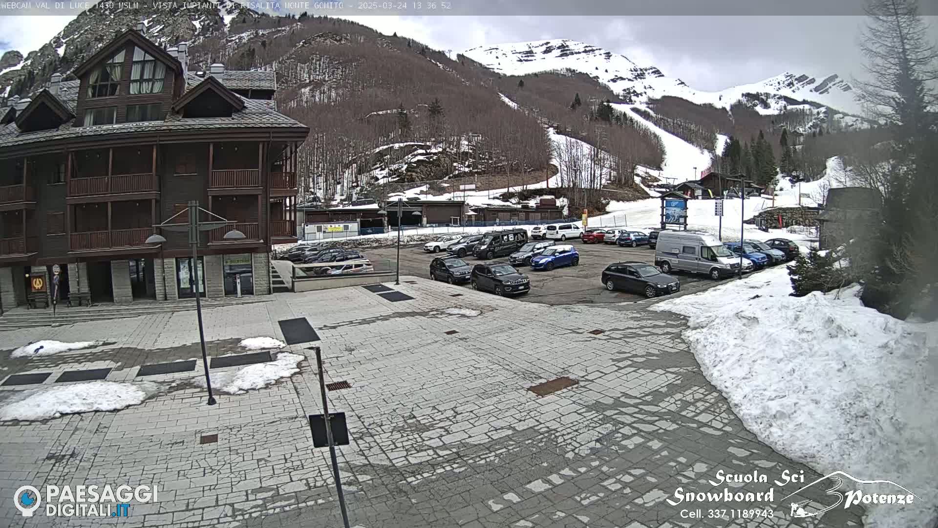 A mostly snow-covered parking lot sits in front of a multi-story brown building, with a snow-dusted mountain and ski slope in the background.