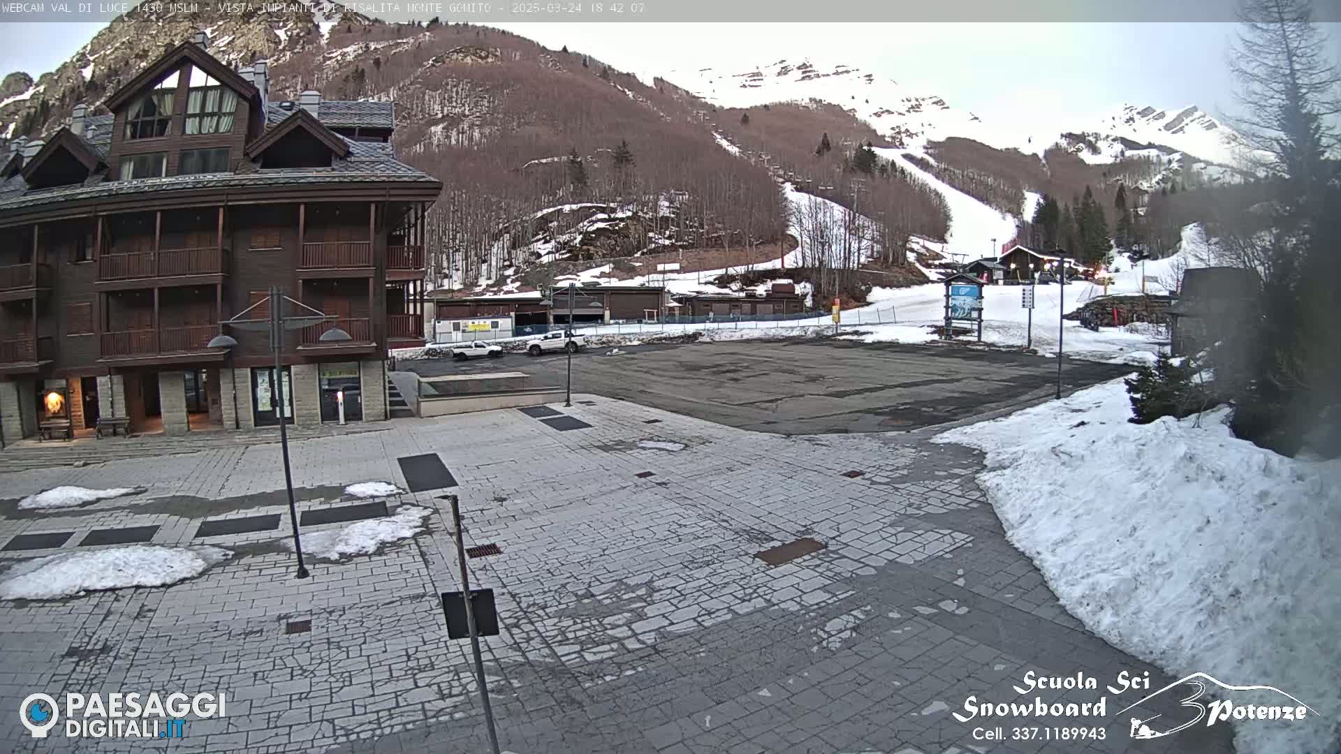 A paved plaza with patches of snow, in front of a multi-story brown building, overlooks a snow-covered mountain with ski slopes.