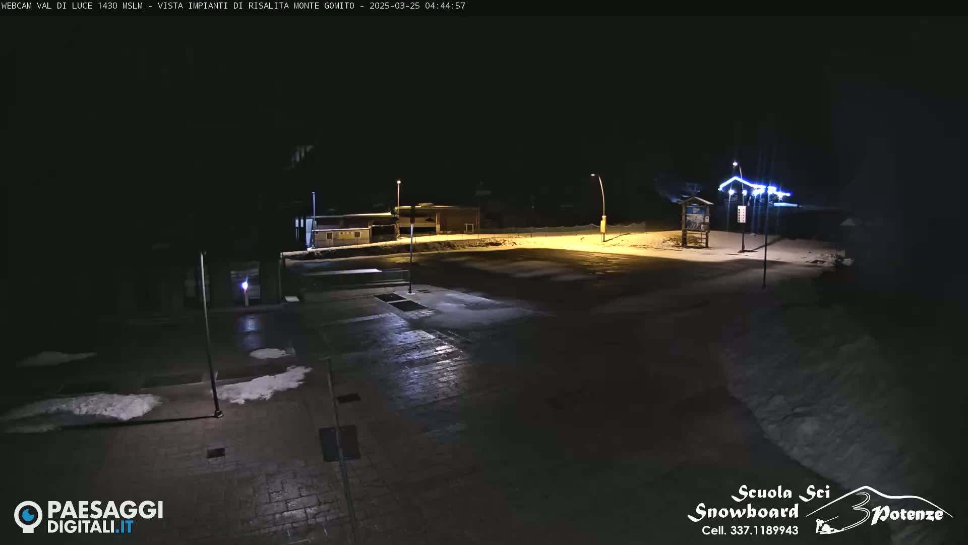 A nighttime view of an empty paved area with patches of snow, illuminated by streetlights, near some buildings.