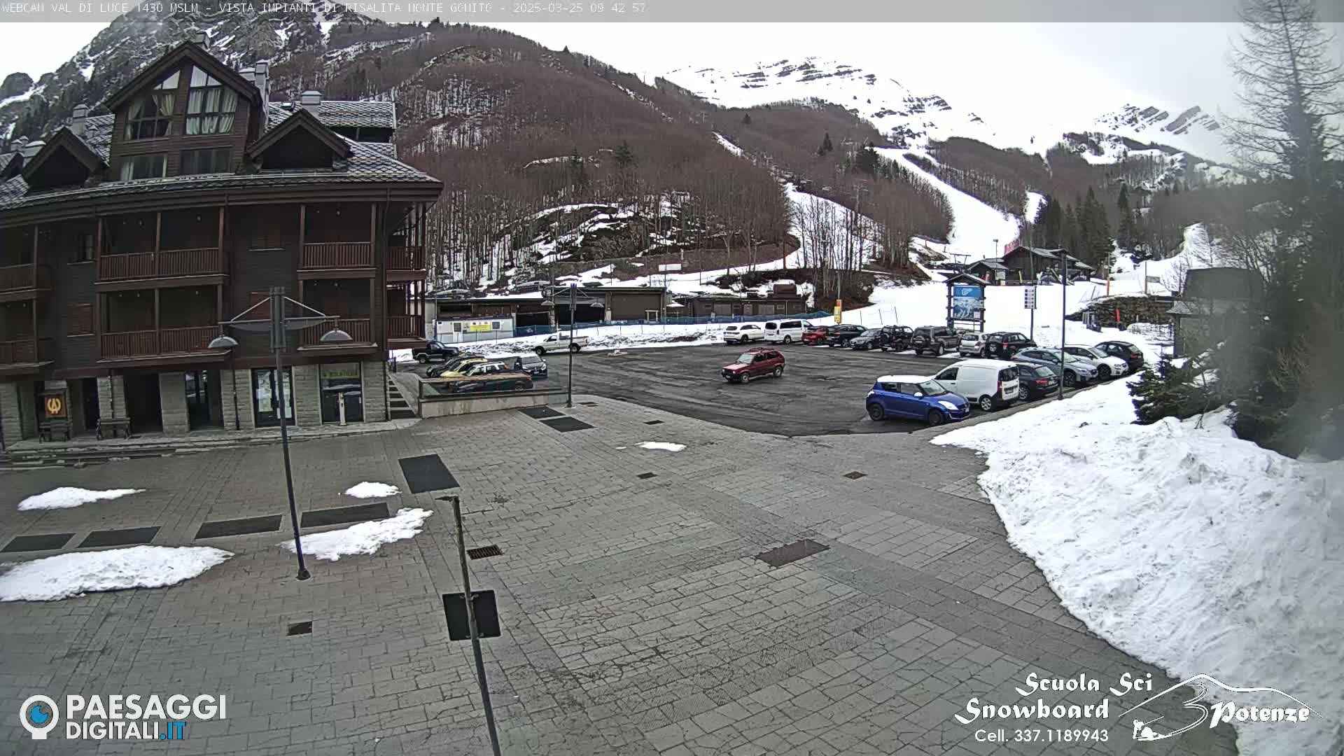 A parking lot with several cars is in front of a multi-story building, with snow patches on the ground and a snow-covered mountain in the background.