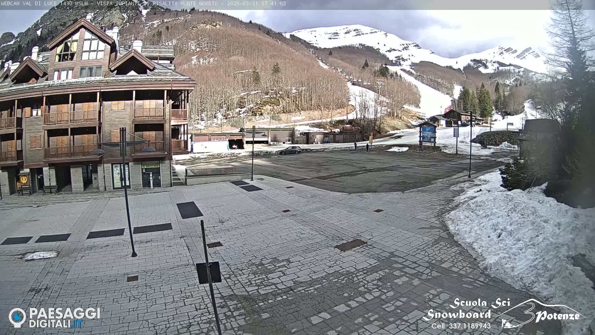 A paved plaza in front of a multi-story building with a mountain in the background showing patches of snow under a partly cloudy sky.
