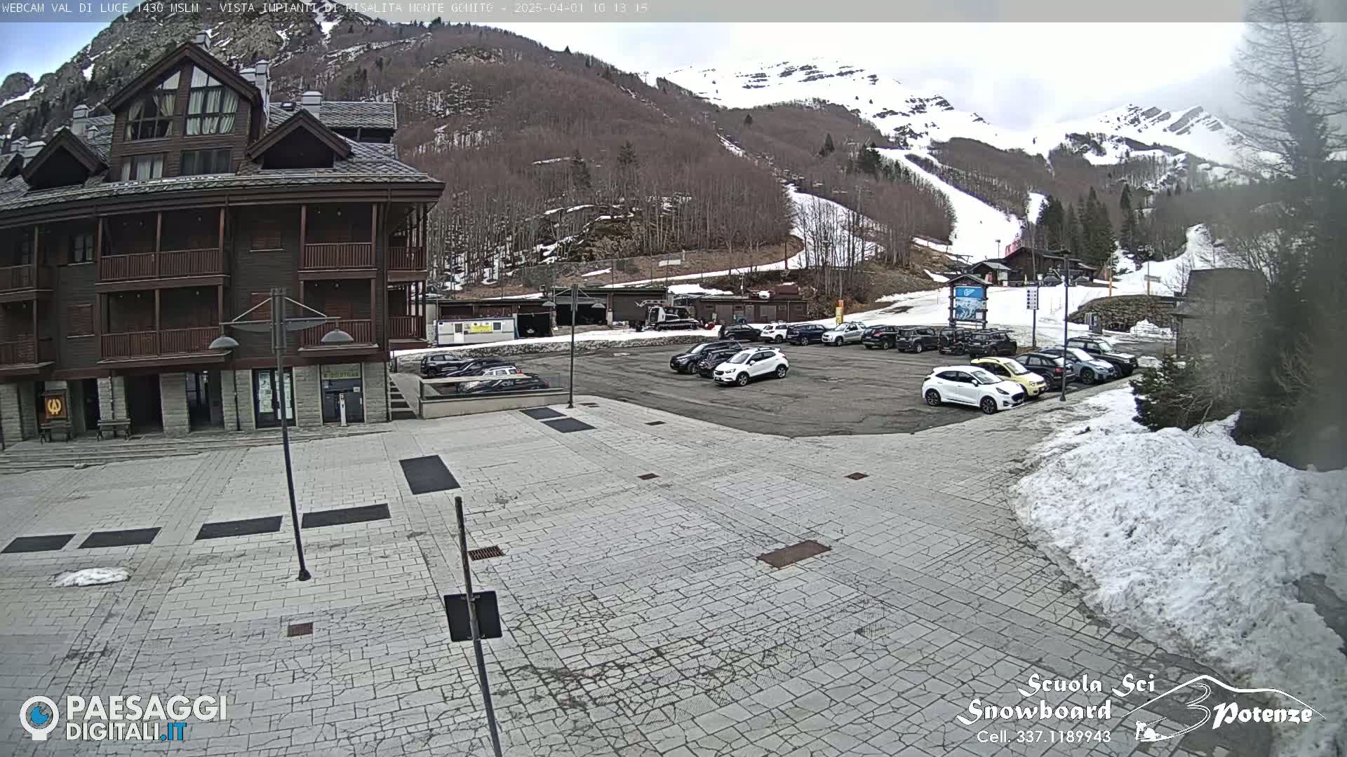 A multi-story brown building with a paved area in front, several cars parked nearby, and a snow-covered mountain in the background under a cloudy sky.