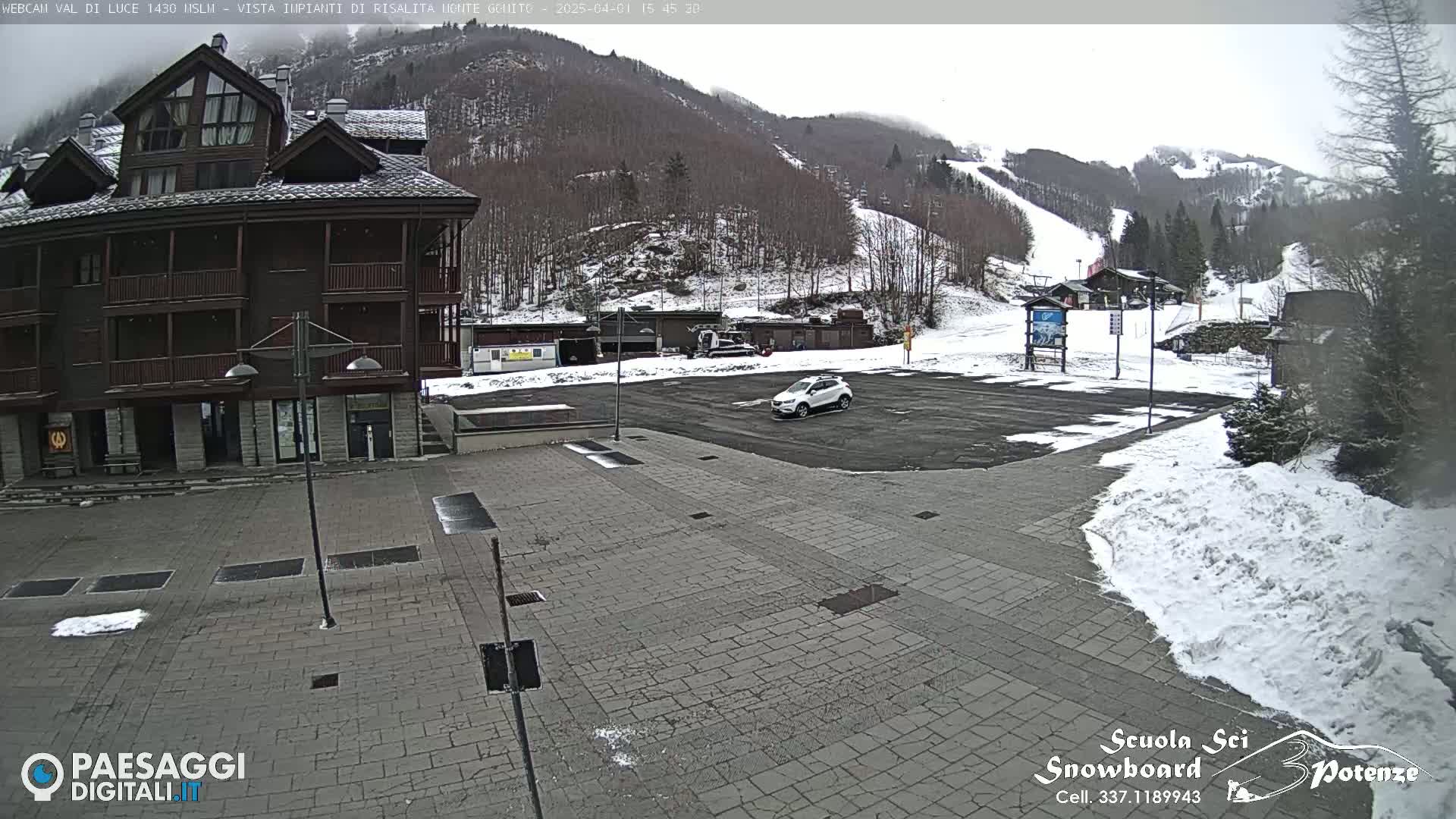 A multi-story brown building sits adjacent to a paved plaza with a car parked in a snowy parking lot overlooking a snow-covered ski slope under an overcast sky.