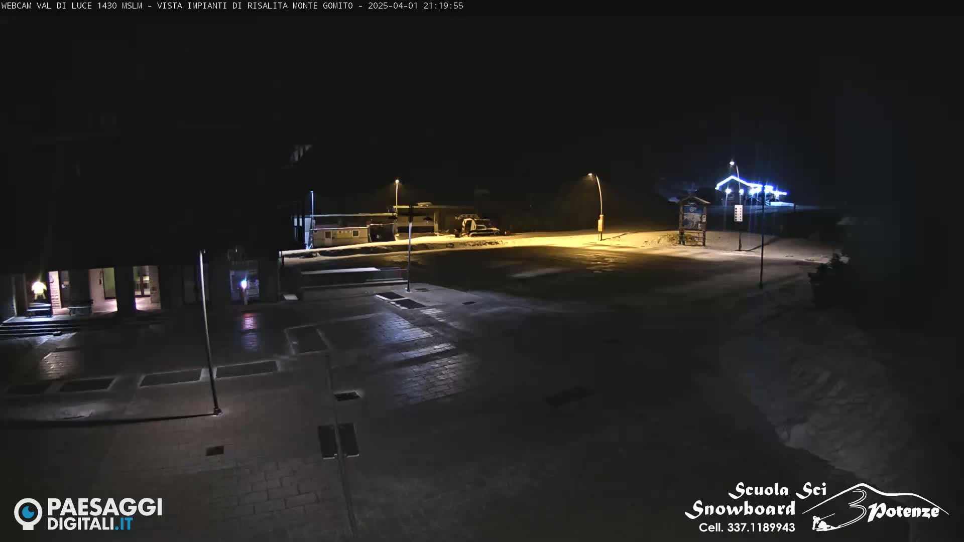 A nighttime, snow-covered parking lot and roadway are illuminated by streetlights.