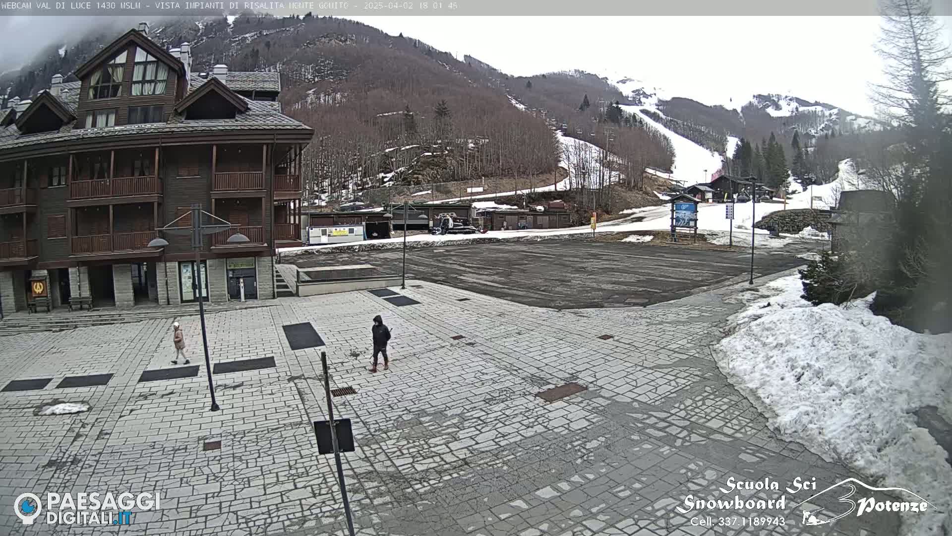 A paved plaza in front of a multi-story building, with a few people walking, overlooks a ski slope on a cloudy day with some snow on the ground.