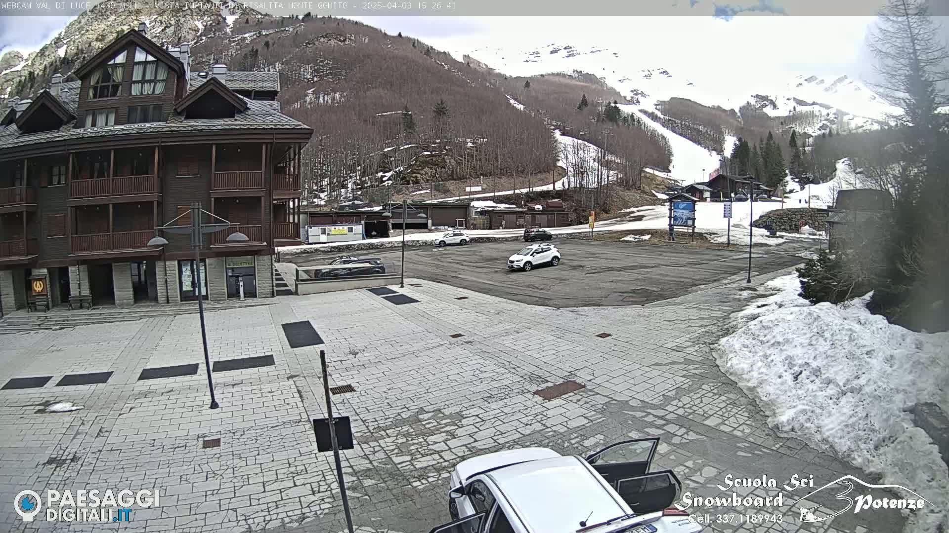 A paved plaza sits in front of a multi-story brown building, with a parking lot and snow-covered mountains in the background under a partly cloudy sky.