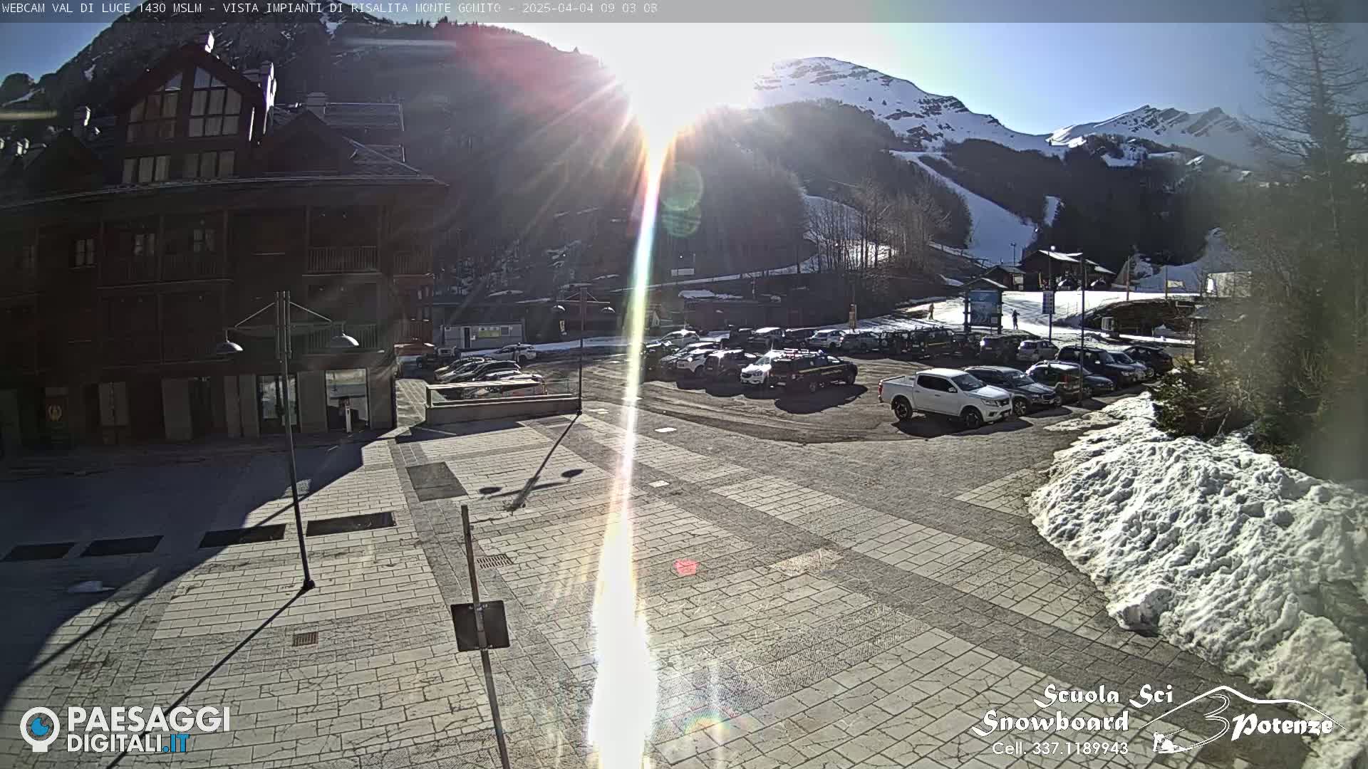 A sunny day in a mountain town shows a paved plaza with parked cars in front of a dark brown building, with snow-covered ground and mountains in the background.