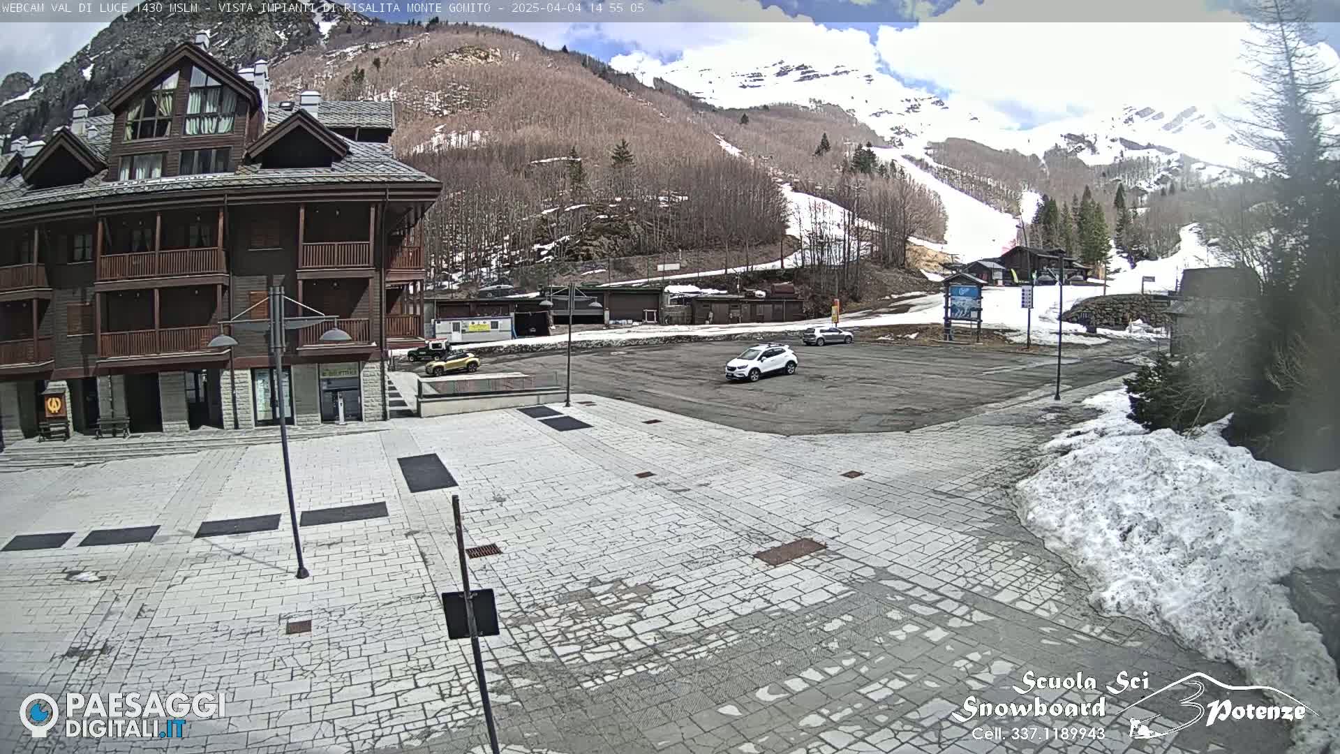 A paved plaza with a few cars in a parking lot sits in front of a multi-story building, overlooking a snow-covered mountain slope under a partly sunny sky.