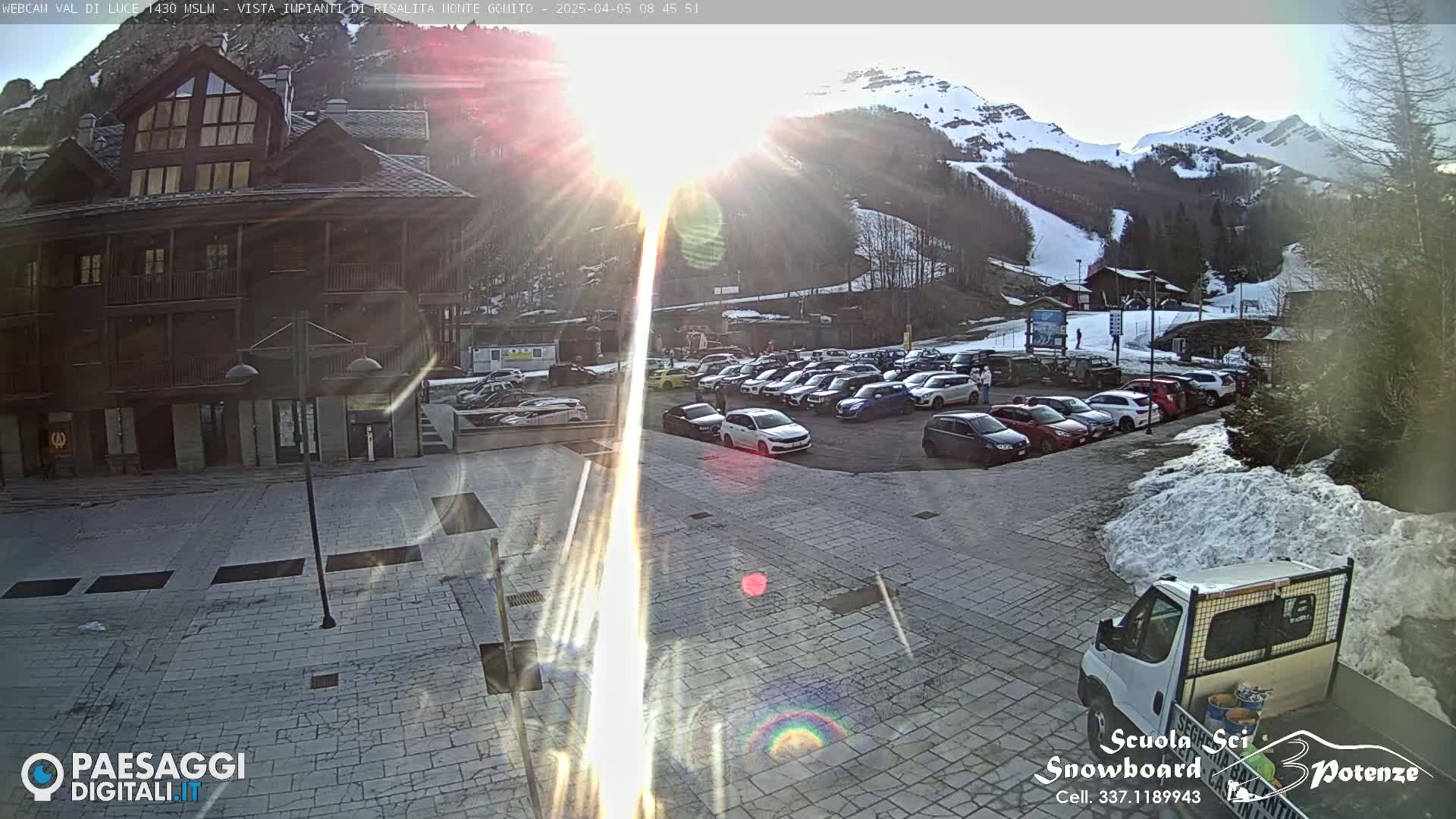 A sunny, snow-covered mountainside overlooks a paved plaza with parked cars and a van, near a multi-story wooden building.
