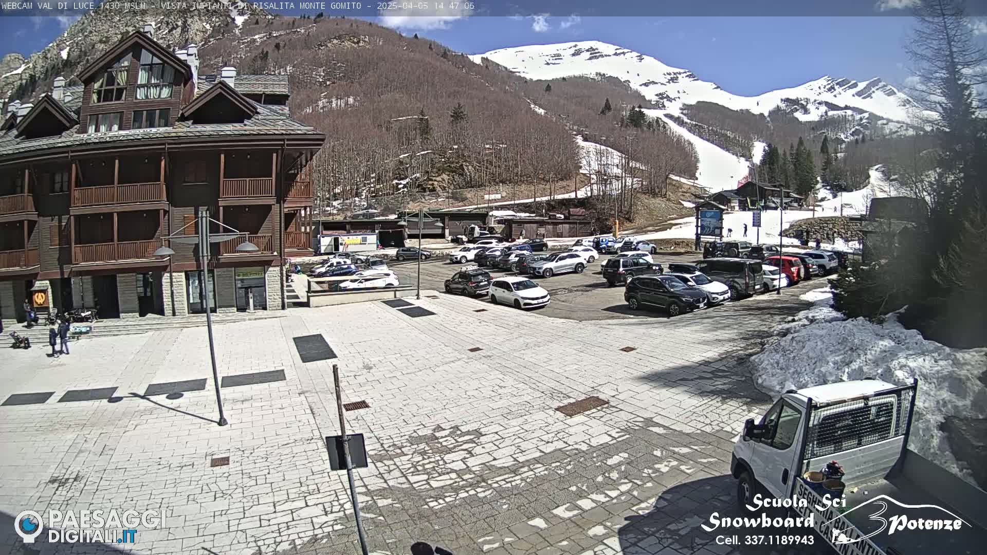 A paved plaza with parked cars sits in front of a multi-story building, overlooking a snow-dusted mountainside under a mostly sunny sky.